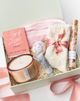 A top view of a luxurious self-care gift set featuring a pink satin sleep mask, a Cup of Love rose tea for two, a 9 oz. Sweet Fig luxury keepsake candle, cloud ivory socks, No. 09 Santal shea butter hand crème, a rose petal bath bomb, and rose bath salts, all arranged in a white gift box with ivory shred and tied with a soft pink ribbon on a white background.