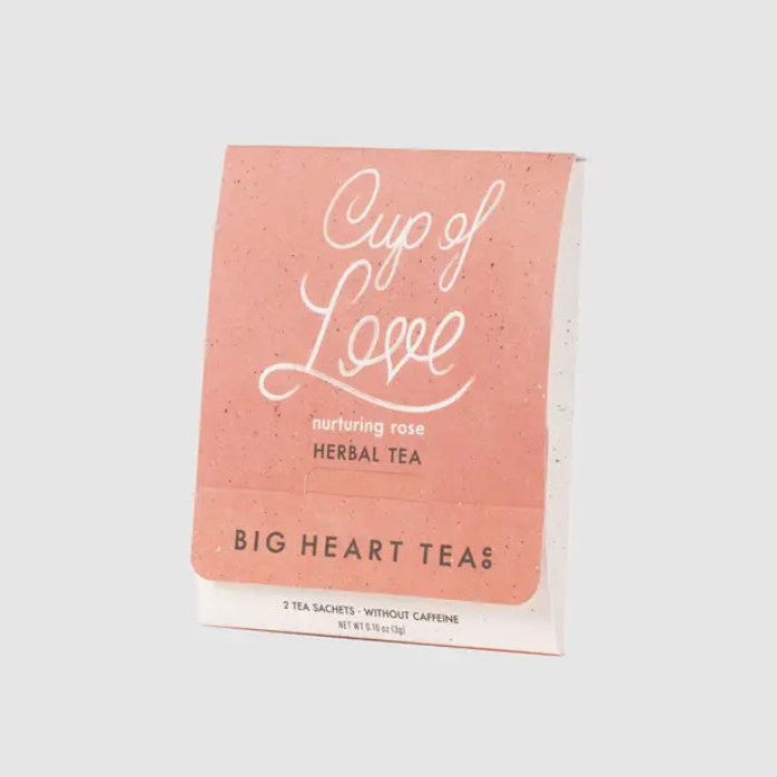 Cup of Love, rose, herbal tea,  the tea package itself, featuring a soft, coral-pink color on the front surface. Large, elegant cursive lettering spells out "Cup of Love" in white, suggesting a comforting and romantic experience. Below that, in a smaller, more straightforward font, are the words "nurturing rose" and "HERBAL TEA." At the bottom, "BIG HEART TEAS" is printed in block capital letters. The package also includes information such as "2 TEA SACHETS - WITHOUT CAFFEINE" and weight.