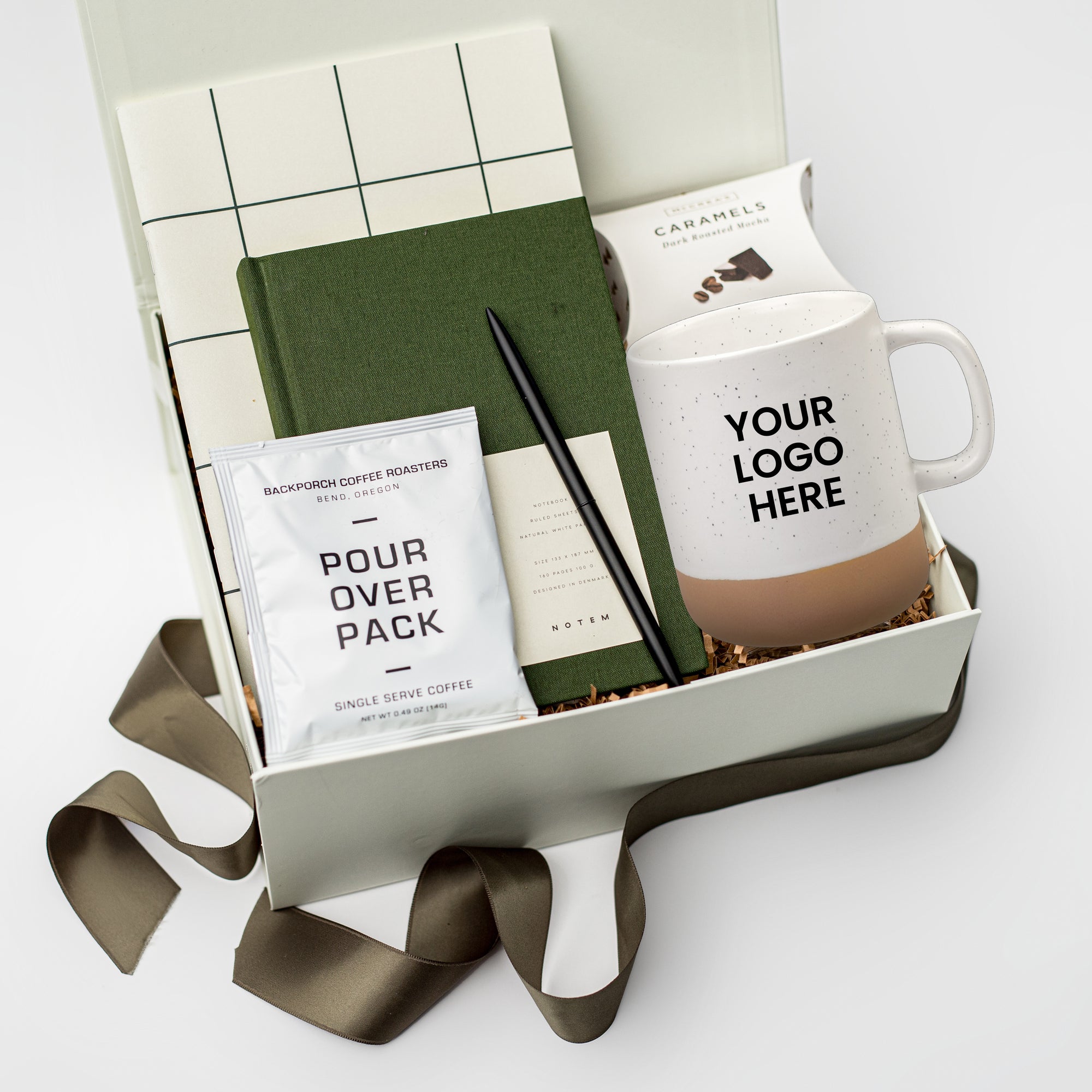 A white gift box filled with brown paper shred and tied with a green ribbon on a white background. Inside the box are a logo tag and logo postcard, a Notem Milo weekly planner, a Notem green linen hardcover notebook, a minimalist black ballpoint pen, a 12oz speckled mug with a custom logo, a single-serve pour-over coffee packet, and handcrafted Cape Cod sea salt caramels — a branded corporate stationery and coffee gift box.