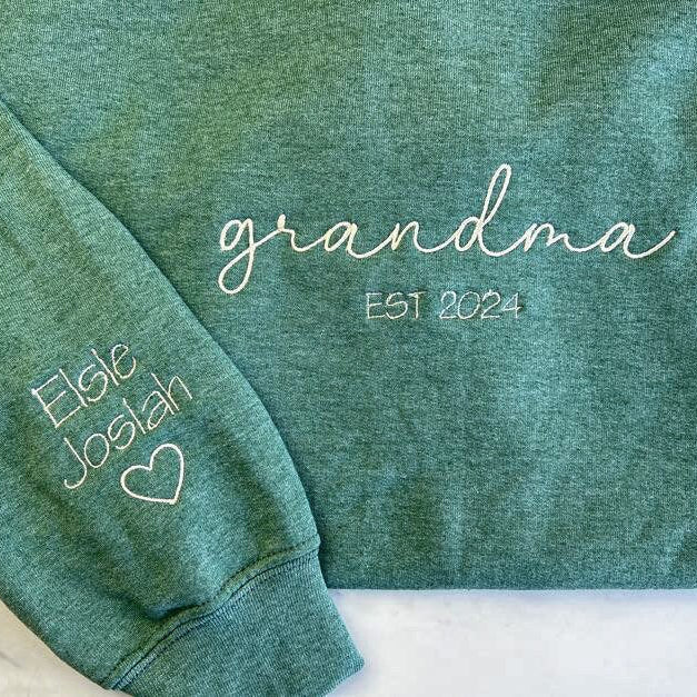 Custom grandma embroidered sweatshirt in color green embroidered in white in the front Grandma Est. 2024 and in the sleeve Elsie and Josiah with a heart emoji on a white background.  