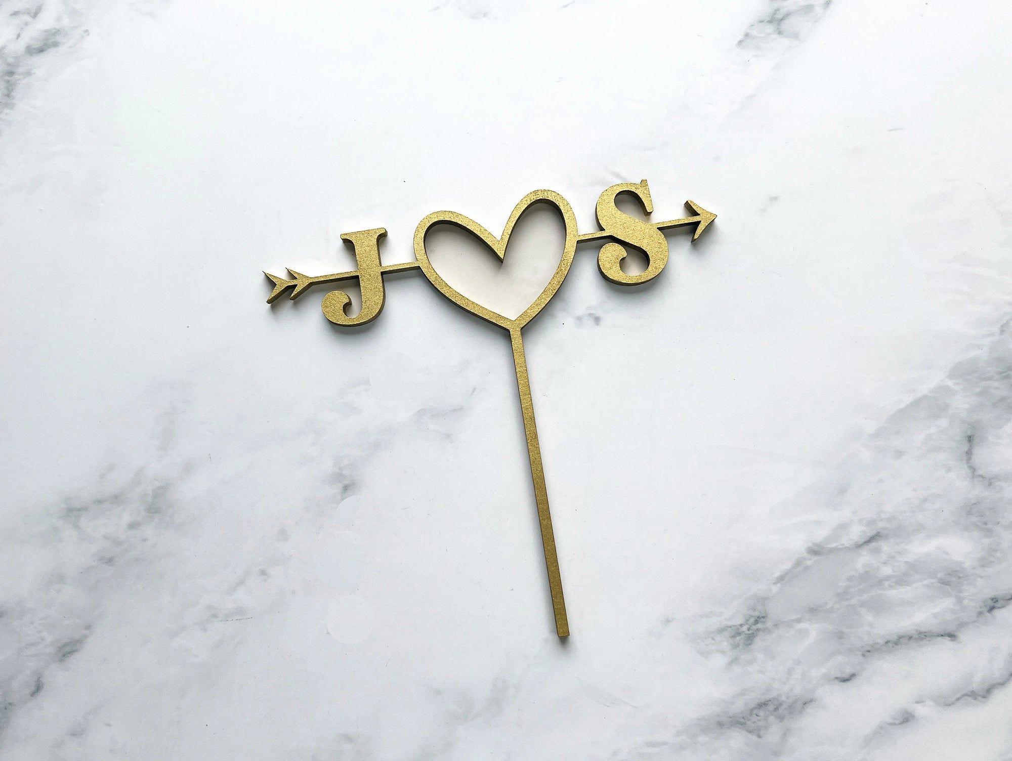 Custom initial wooden cake topper is featured with the initials J heart S with an arrow across in gold color in a marble like background. 