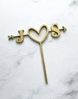 Custom initial wooden cake topper is featured with the initials J heart S with an arrow across in gold color in a marble like background. 