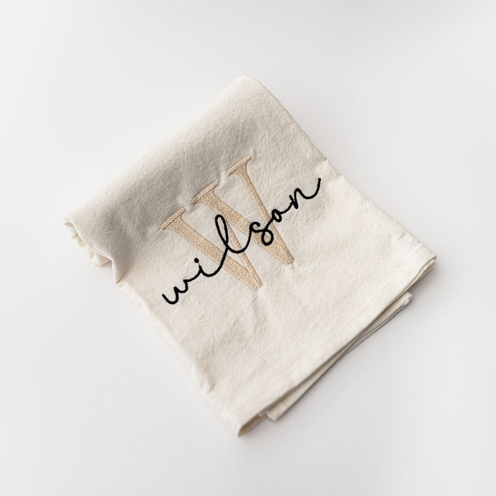 Custom name embroidered white tea towel with W in champagne color and Wilson in black lettering on a white background. 