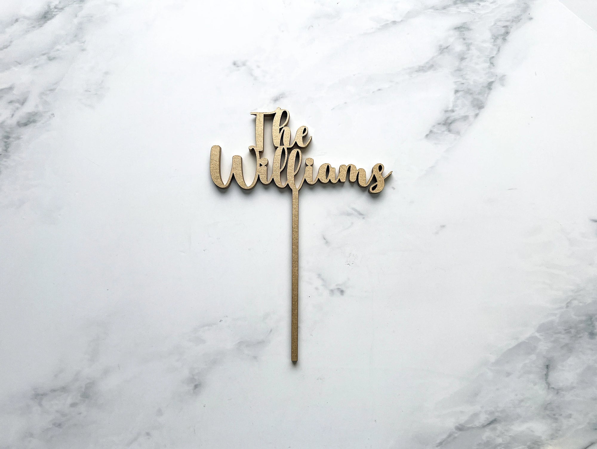 Custom name wooden cake topper is featured in champagne color personalized "The Williams" on a marble like background. 