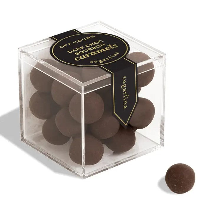 Dark chocolate bourbon caramels from Sugarfina are featured in their square shaped transparent container displaying the caramels inside with black label and gold lettering one of them is displayed out of the box round and dark brown colored on a white background. 