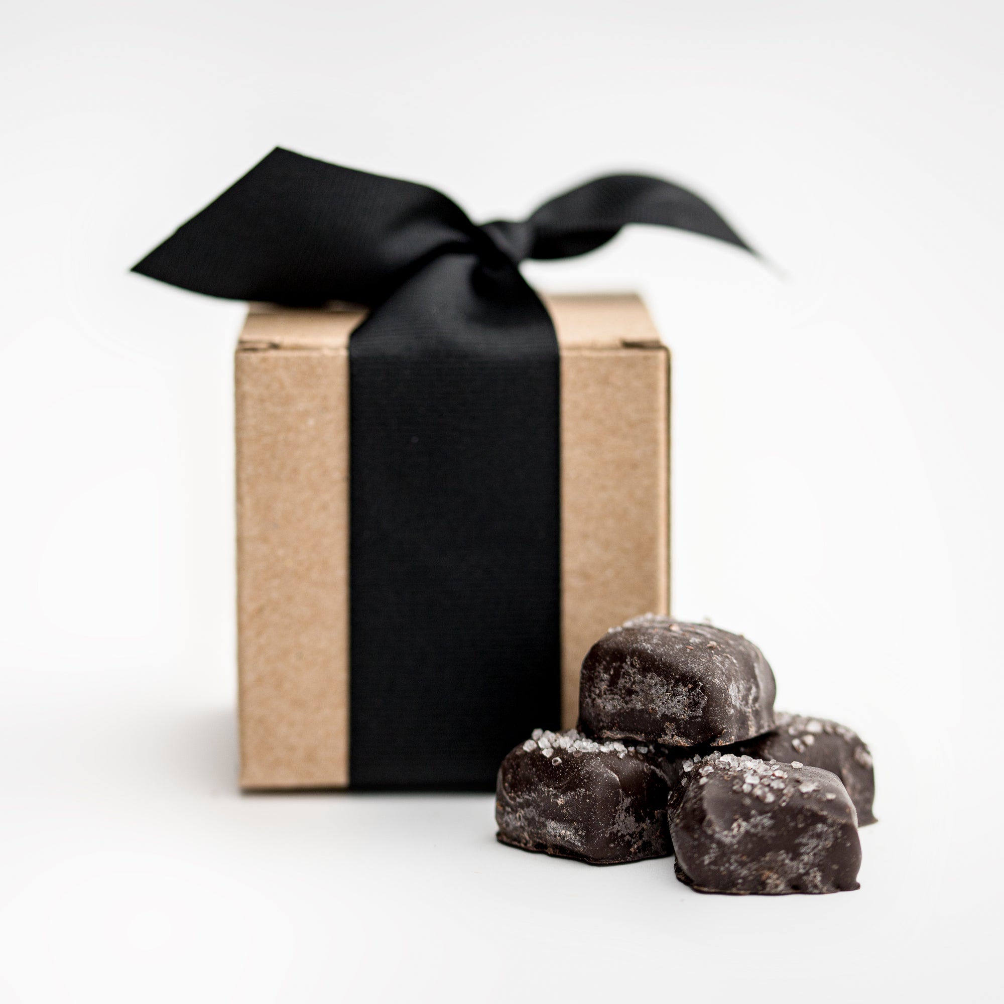 Dark chocolate sea salt covered caramels featured in front 4 dark chocolates with sea salt and in the back of them the card stock light brown gift box they come in wrapped in a black ribbon. 
