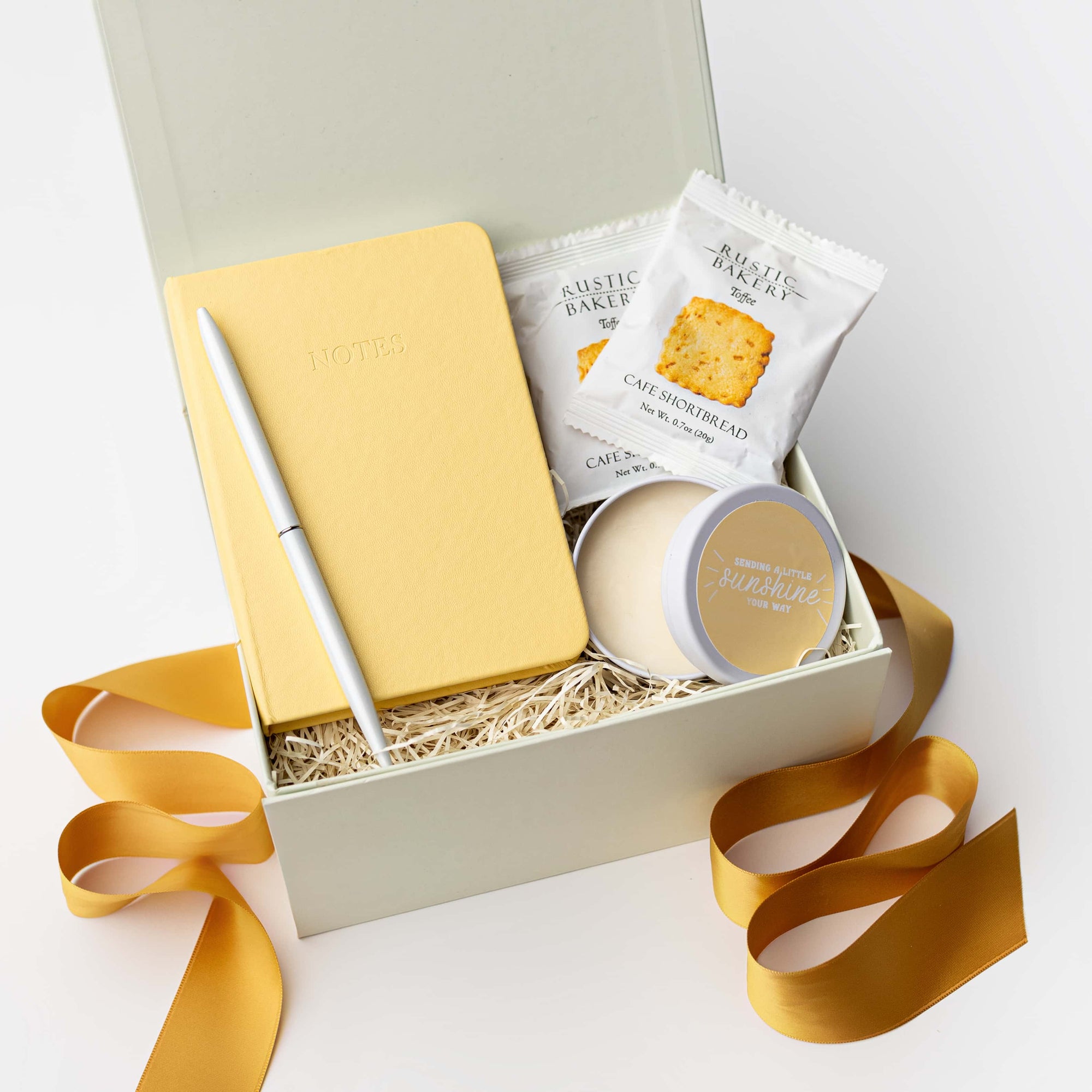 The Day Brightener Gift Box is featured in our signature white gift box filled with ivory shred, containing, from left to right, a yellow softcover notebook, a white minimalist bullet pen, two toffee shortbread cookies, and a “Sending Sunshine” candle. Displayed on a white background, surrounded by an orange ribbon.