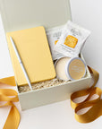 The Day Brightener Gift Box is featured in our signature white gift box filled with ivory shred, containing, from left to right, a yellow softcover notebook, a white minimalist bullet pen, two toffee shortbread cookies, and a “Sending Sunshine” candle. Displayed on a white background, surrounded by an orange ribbon.