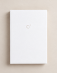 A minimalist white hardcover notebook or journal with a small gold circular logo on the front cover, centered on a warm beige background.