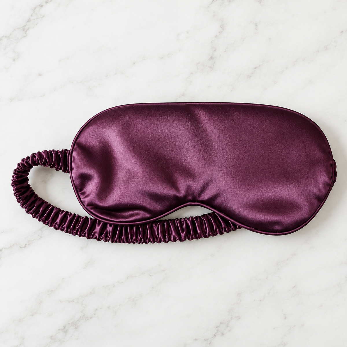 A satin sleep mask in a deep plum purple color, featuring an elasticized ruffled strap, laid flat on a white marble background.
