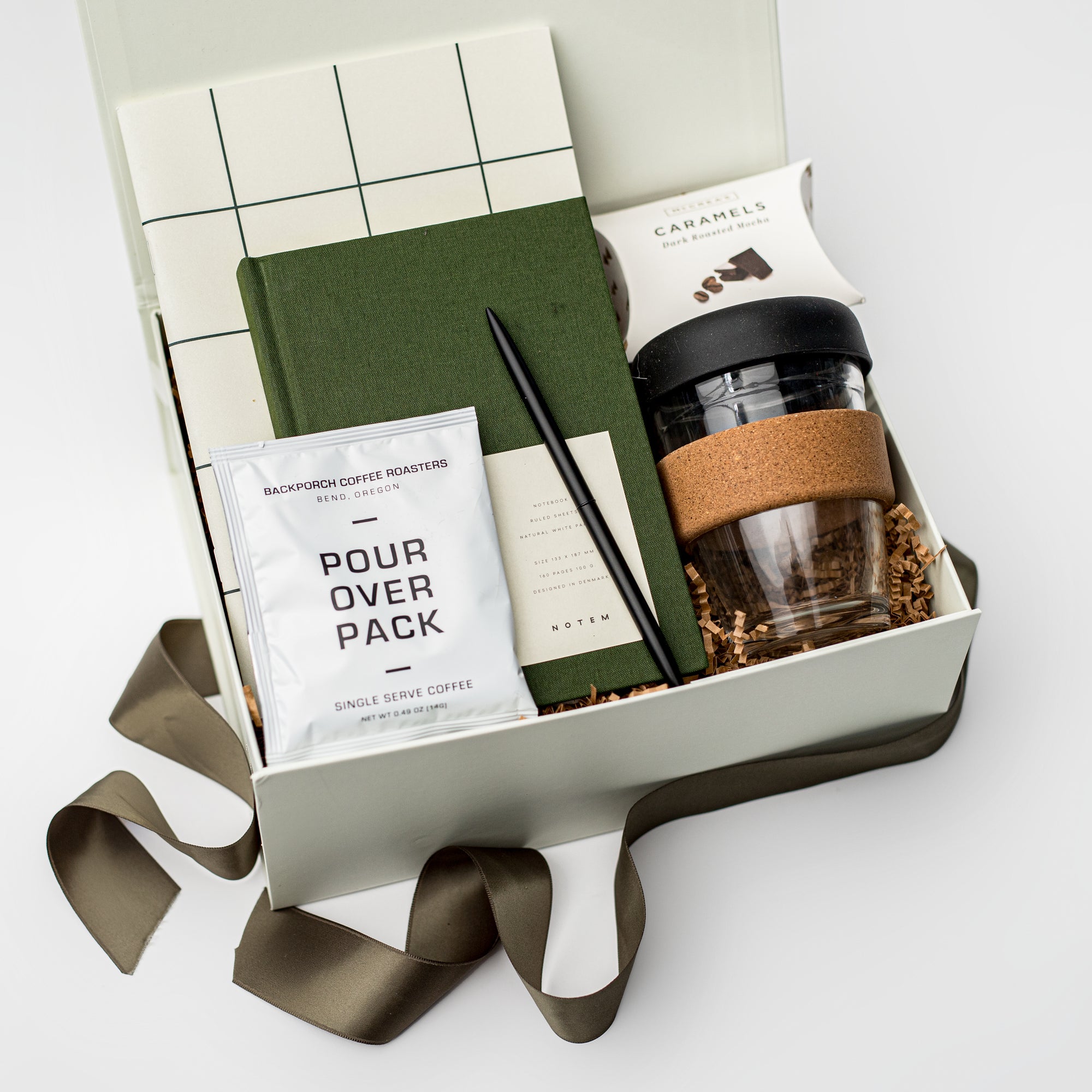 The Office Essentials New Hire Curated Gift Box is featured in our signature white box filled with brown shredded paper, containing, from left to right, a Notem Milo weekly planner with a green linen cover, a minimalist black ballpoint pen, a single pour-over coffee packet, Cape Cod sea salt caramels, and a glass travel mug with a cork sleeve.
