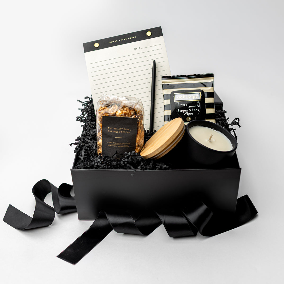 A curated gift box set in a matte black box with black shredded paper filler and ribbon, containing a candle, a notebook, a bag of caramel corn, and a small appliance, photographed on a clean light gray studio background.