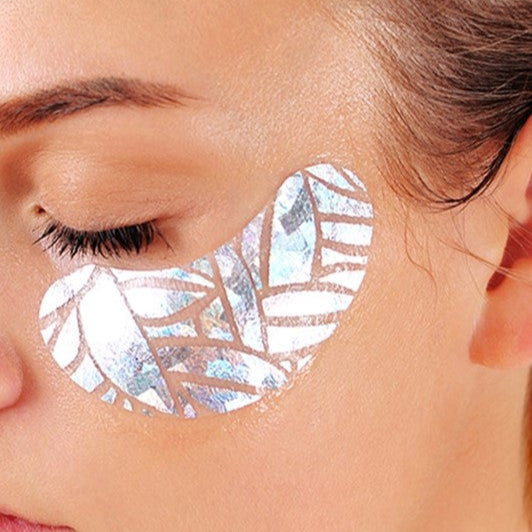 close-up view of a person's eye area with a decorative, iridescent eye patch applied beneath the closed eyelid.

Focused on the upper portion of a person's face, specifically highlighting the eye and the surrounding skin. The eye is closed, with eyelashes visible. Below the eye, a patch is applied; it is an abstract design with a light-reflective, shimmering surface. The frame is tight, centered on the eye area, allowing for a detailed view of skin texture and the texture of the patch. 