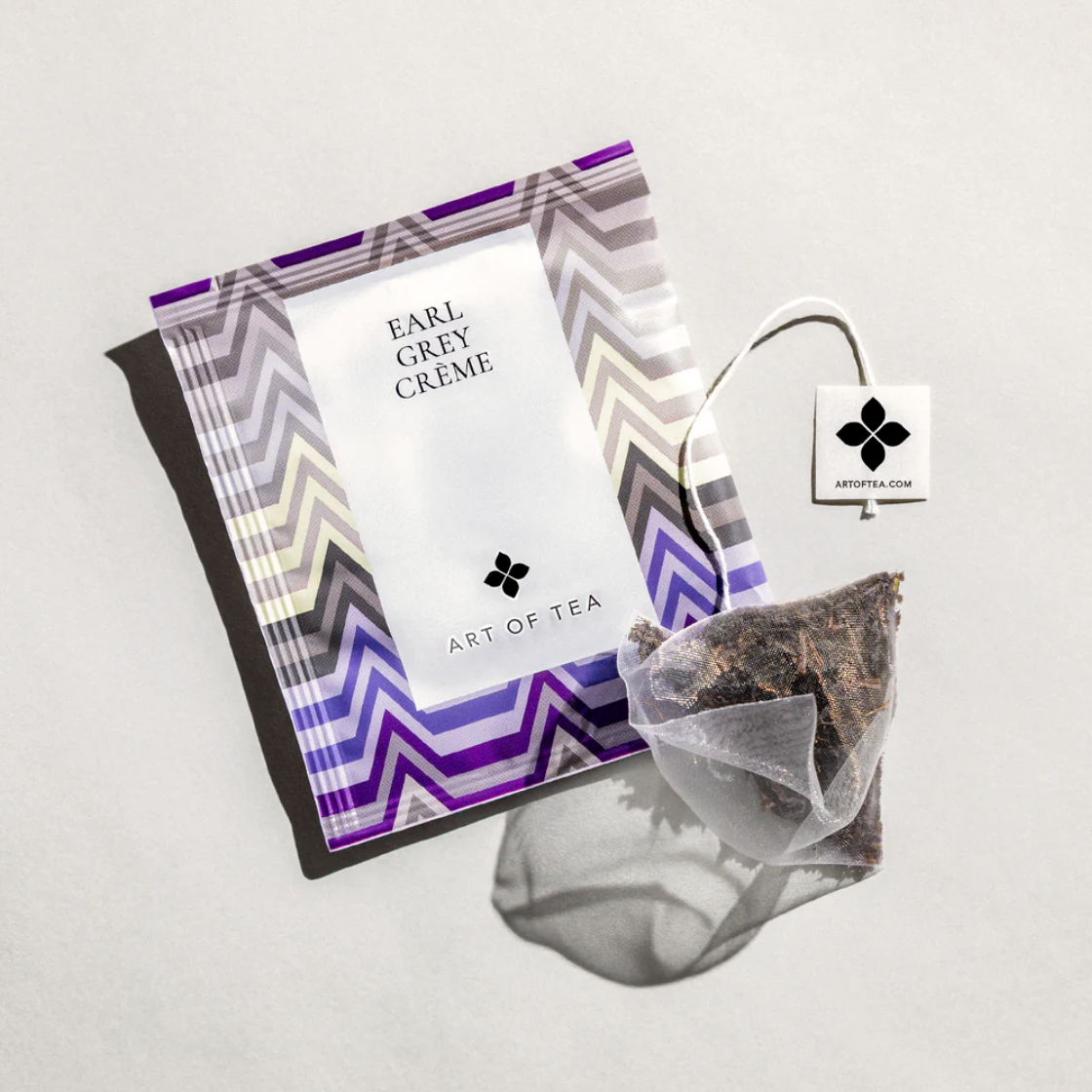 Earl grey creme sachets is featured in its square shaped sachet with purple grey lines white label with black lettering in front of it the tea bag itself out of its container. on a white background. 