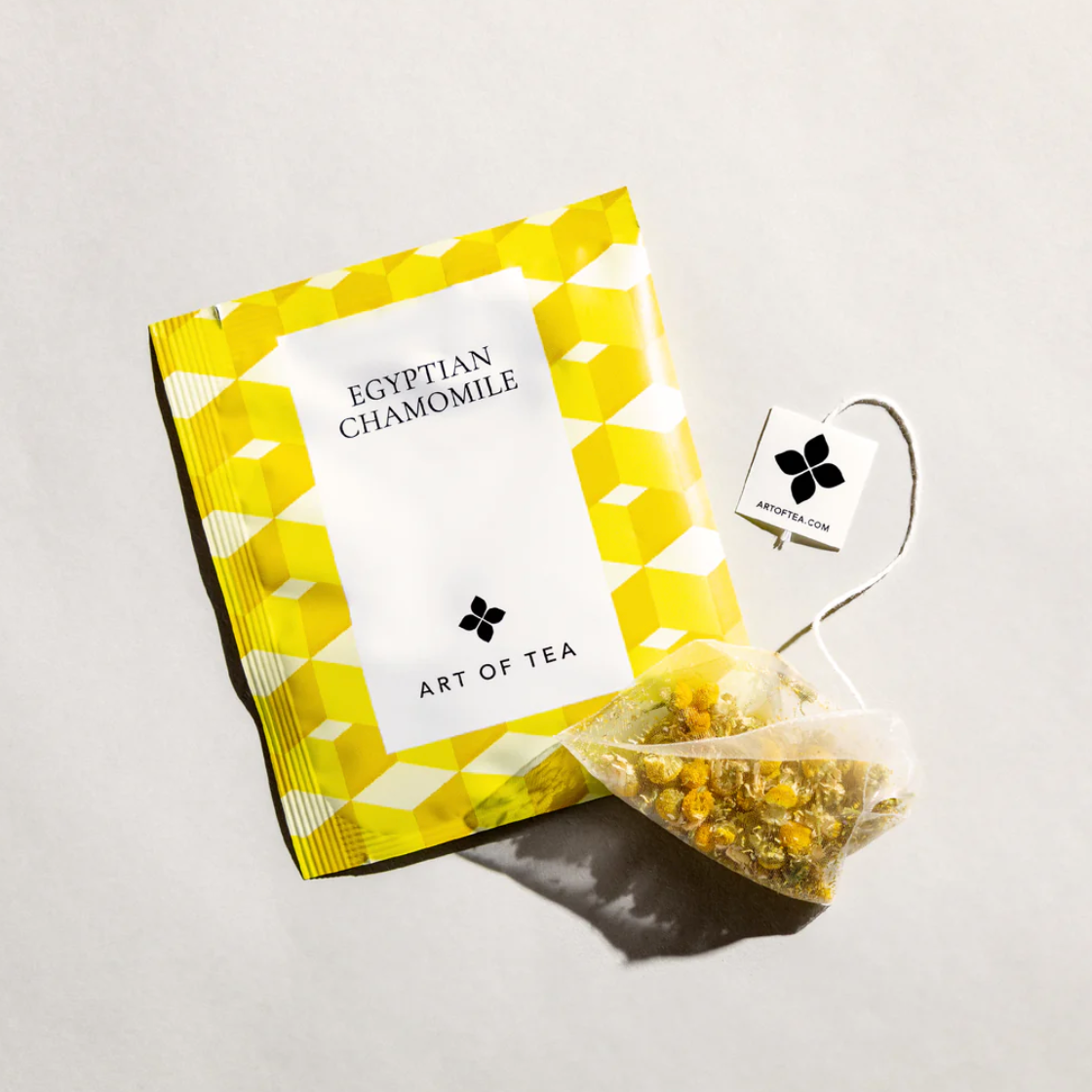 Egyptian chamomile tea sachet is featured in its square shape sachet with yellow hexagons white label with black lettering and in front of the sachet the tea itself out of its package on a white background. 