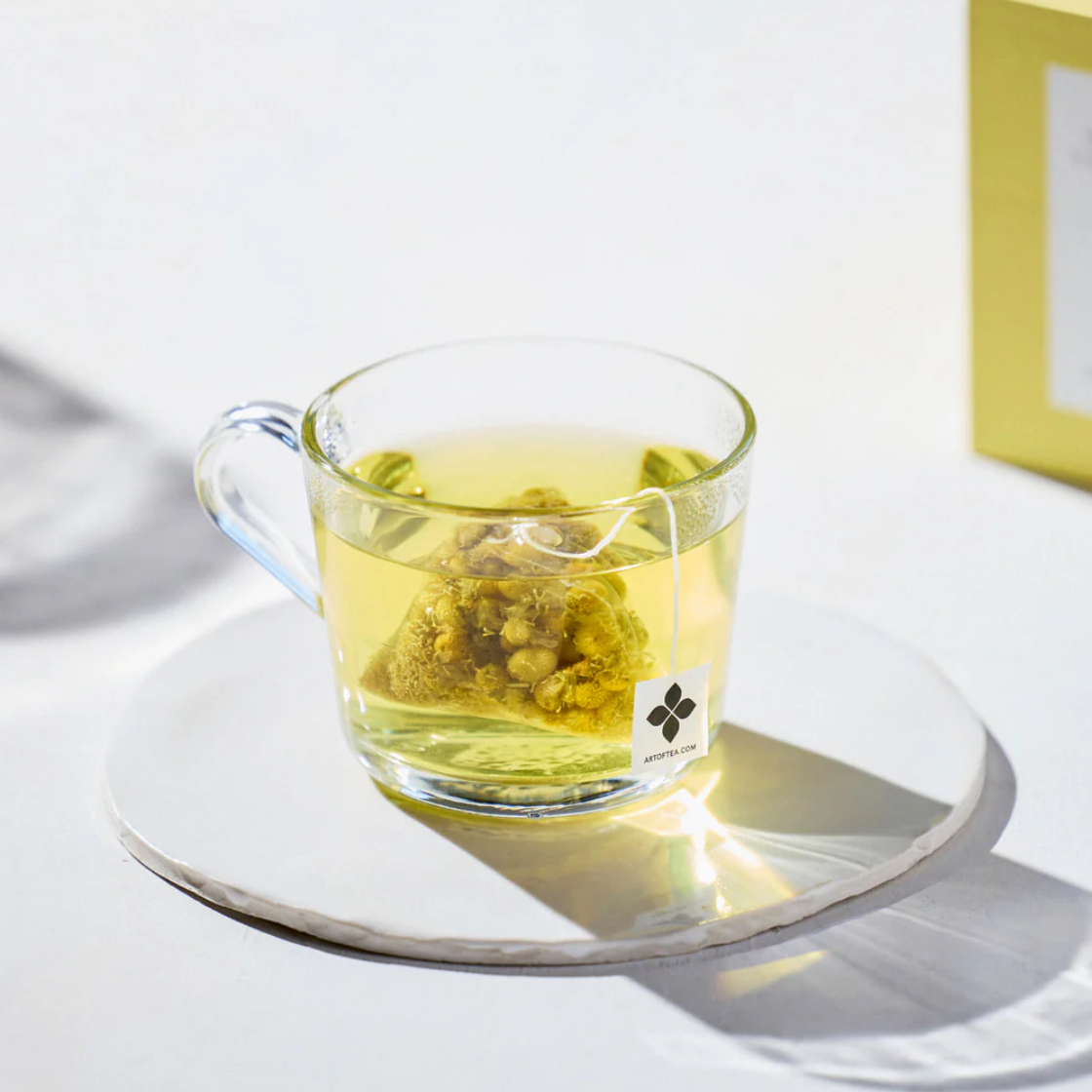 Egyptian chamomile tea sachet featured in a clear glass mug showcasing the tea bag inside that gives the water a warm yellow color on a white background. 