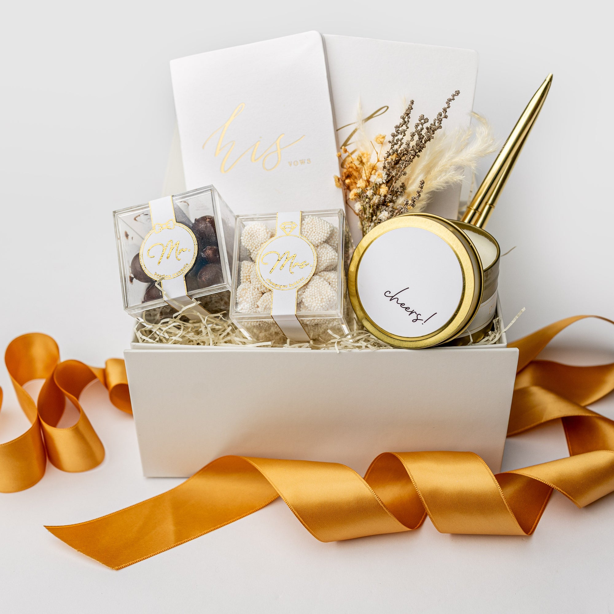 Engagement celebration gift box displayed on a white background with ivory shred and an orange ribbon, containing a set of foil-stamped vow books, two gold minimalist bullet pens, Mrs. champagne bubbles gummies, Mr. cordials caramels, a 4oz hand-poured “Congrats” candle, and a dried floral mini bouquet.