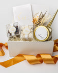 Engagement celebration gift box displayed on a white background with ivory shred and an orange ribbon, containing a set of foil-stamped vow books, two gold minimalist bullet pens, Mrs. champagne bubbles gummies, Mr. cordials caramels, a 4oz hand-poured “Congrats” candle, and a dried floral mini bouquet.
