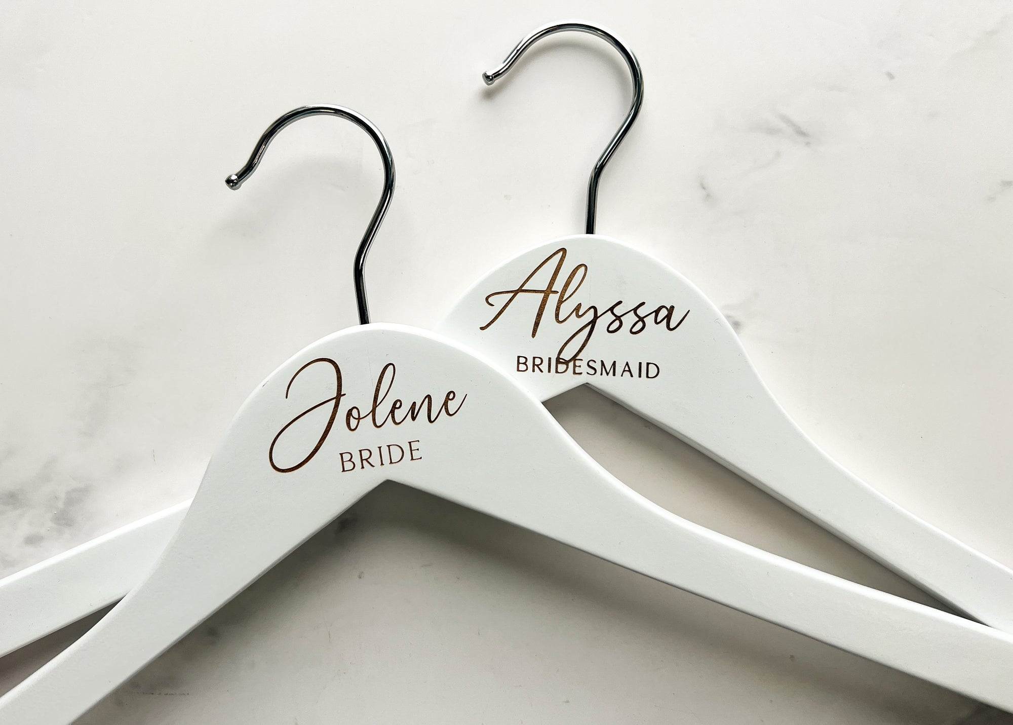 Engraved bride hangers are featured, two white hangers with silver hooks personalized  Jolene -Bride and Alyssa - Bridesmaid on a marble-like background.
