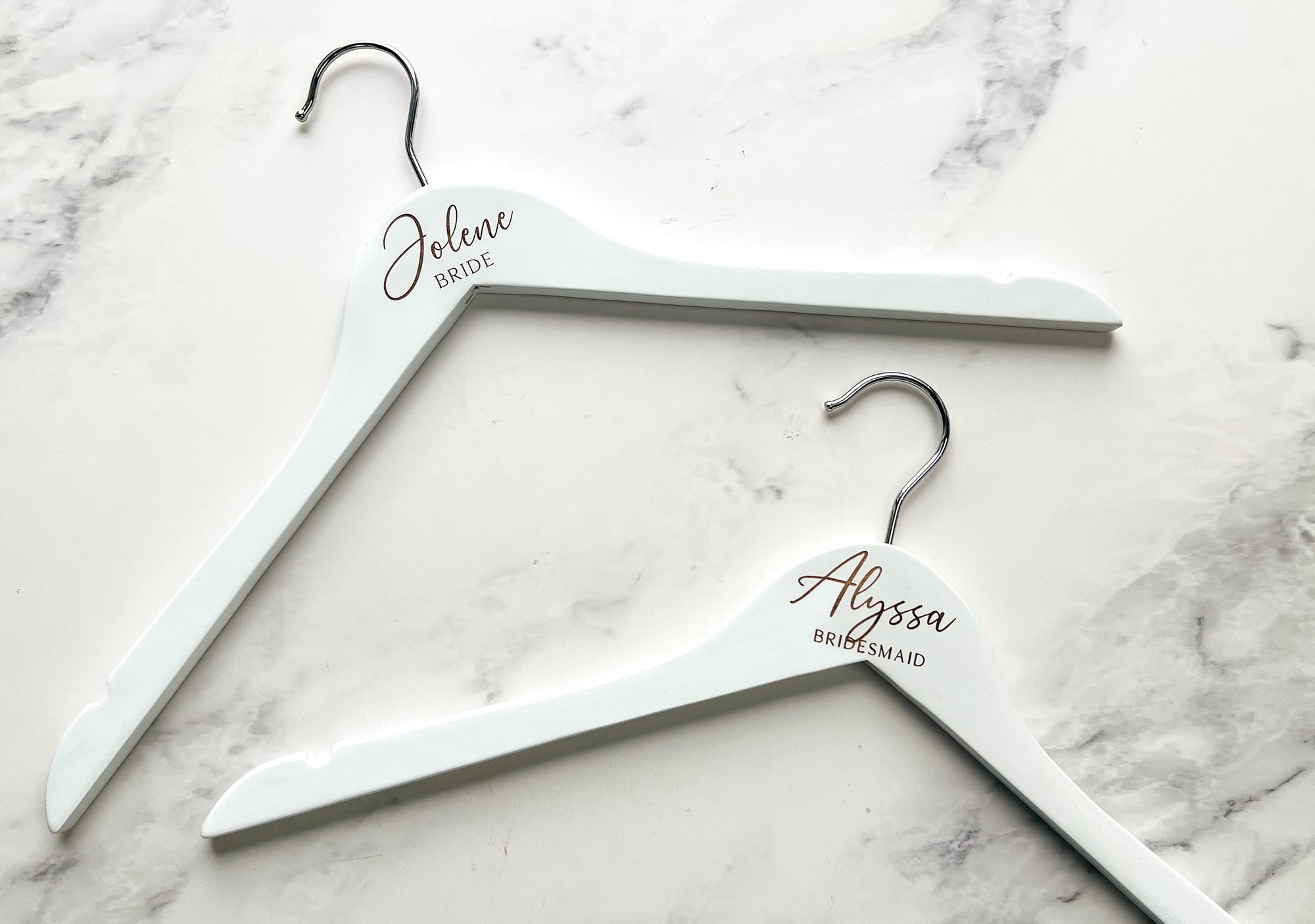 Engraved bride white hangers personalized with Jolene Bride and Alyssa bridesmaid on a marble-like background.