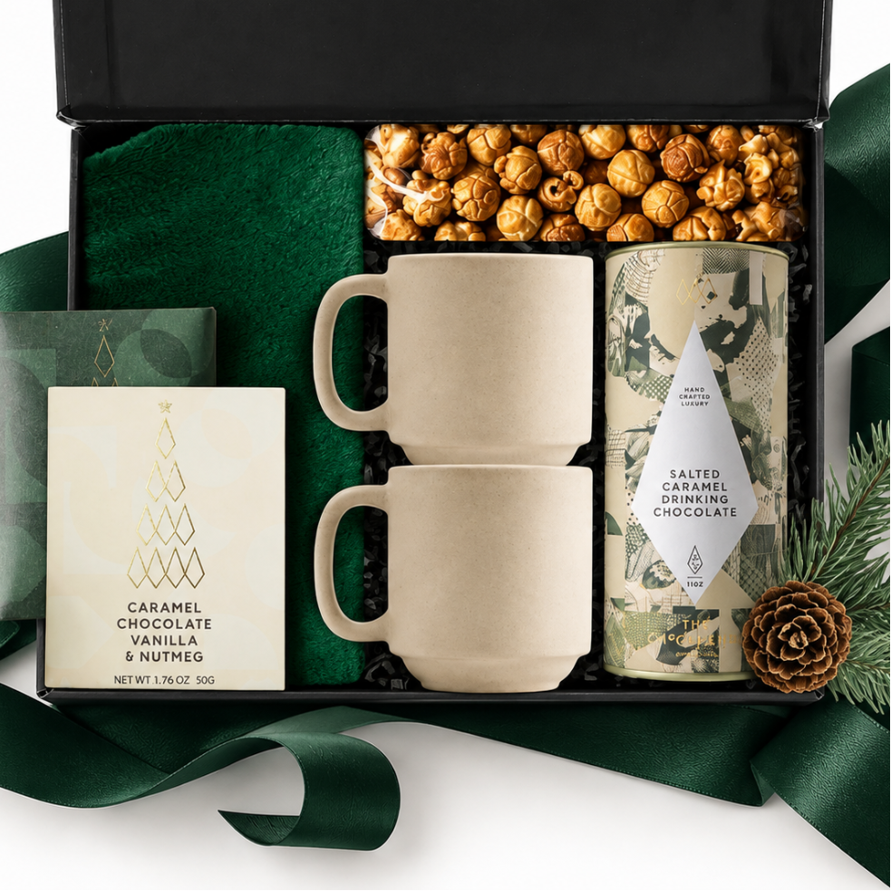 A curated gift box set shown open on a white background, with a black presentation box containing two cream ceramic mugs, a tin of caramel chocolate, a card, a bowl of popcorn, green fabric lining, and dark green ribbon. A pine cone and evergreen sprigs sit beside the box.