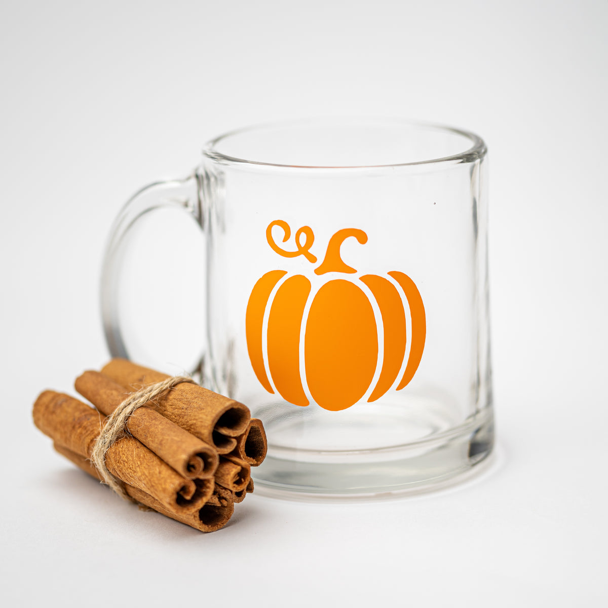 A clear glass mug with a bright orange pumpkin graphic on the front, styled next to a small bundle of cinnamon sticks on a clean white background.