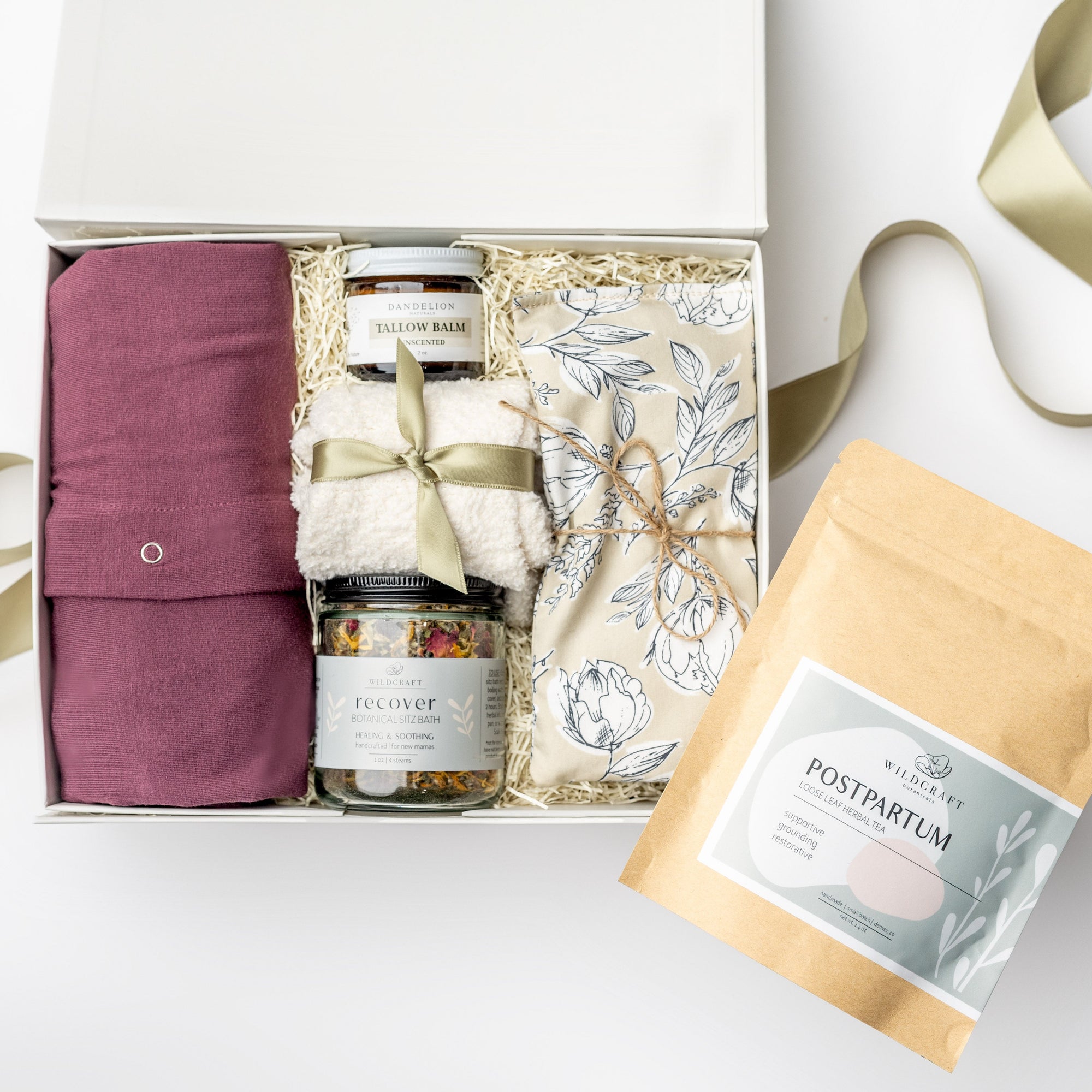 A top view of a postpartum care gift set featuring a cotton nursing gown, Dandelion Tallow Balm, Wildcraft postpartum tea, Wildcraft Recover sitz bath, cozy socks, and a heat pad, all arranged in a white gift box with ivory shred and accented with a green ribbon on a white background.
