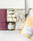 A top view of a postpartum care gift set featuring a cotton nursing gown, Dandelion Tallow Balm, Wildcraft postpartum tea, Wildcraft Recover sitz bath, cozy socks, and a heat pad, all arranged in a white gift box with ivory shred and accented with a green ribbon on a white background.