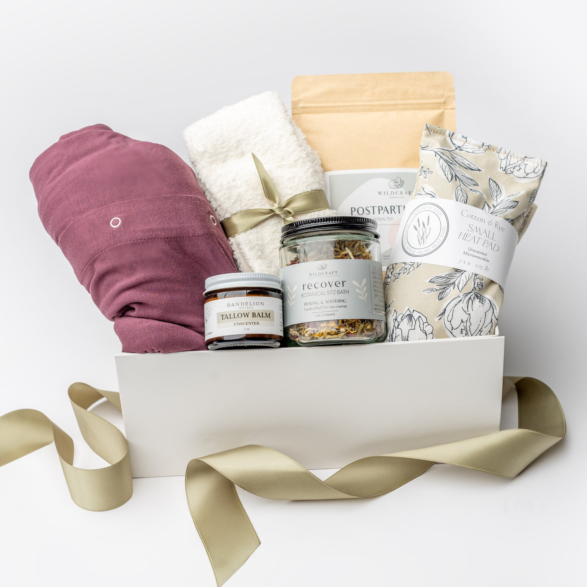A front view of a postpartum care gift set featuring a cotton nursing gown, Dandelion Tallow Balm, Wildcraft postpartum tea, Wildcraft Recover sitz bath, cozy socks, and a heat pad, all arranged in a white gift box with ivory shred and accented with a green ribbon on a white background.