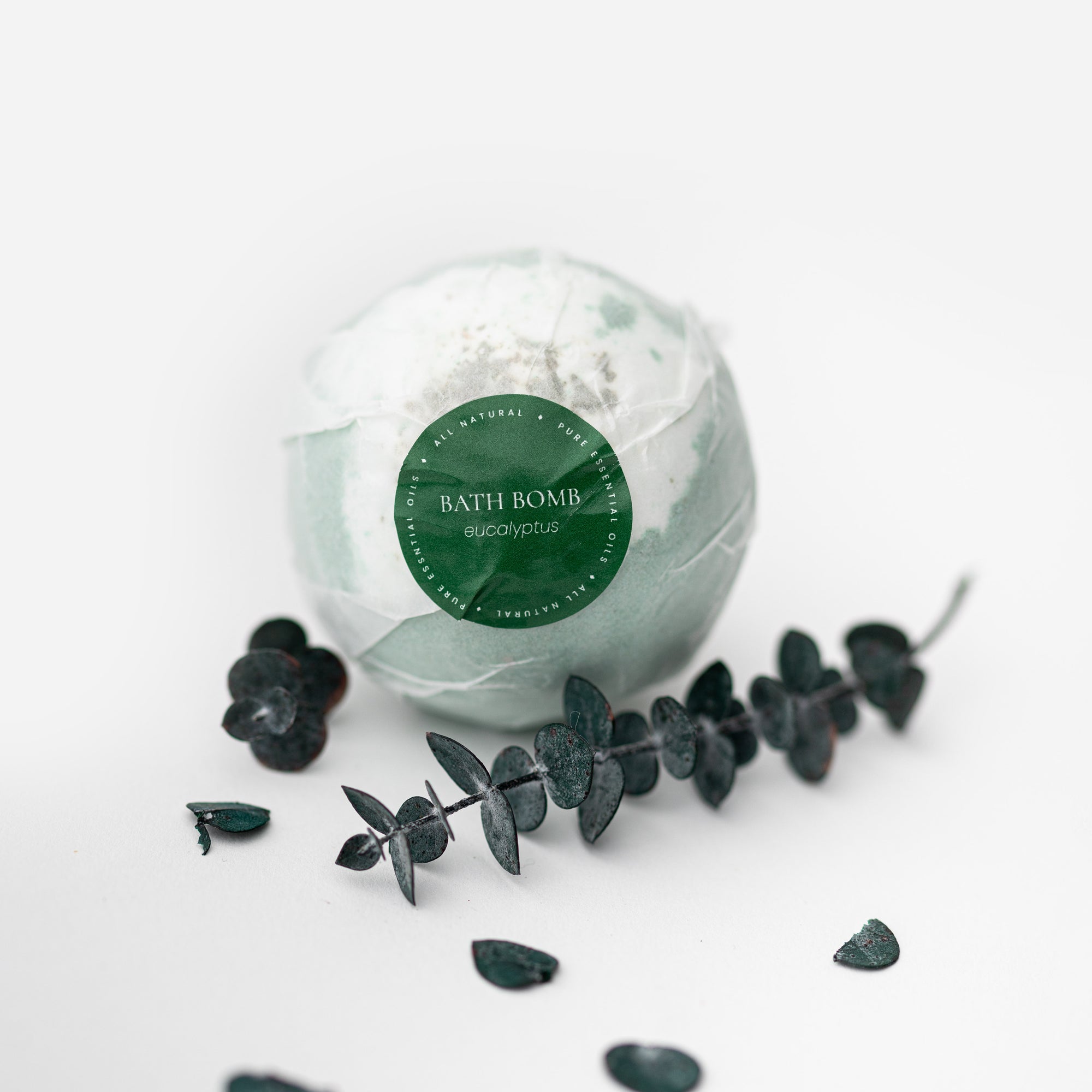 Eucalyptus bath bomb is featured in its white transparent wrapper with green label that has white lettering with some eucalyptus around it. 
