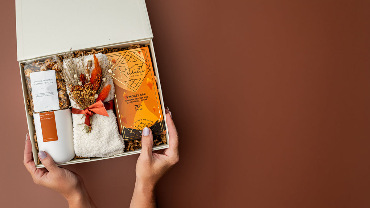 A gift box held by two hands against a solid brown background, containing a white candle, a small dried flower arrangement tied with an orange ribbon, and an orange book or booklet.
