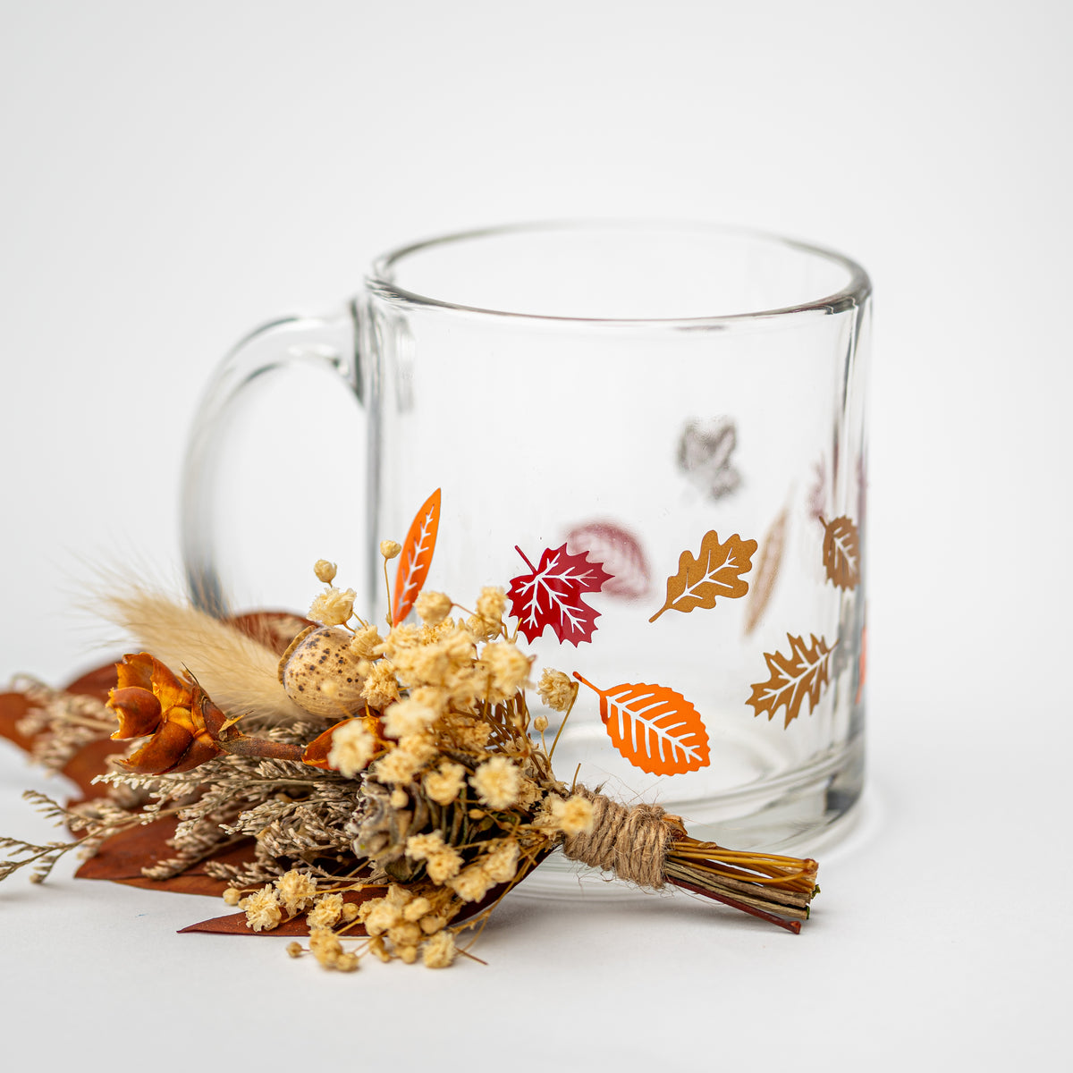 A clear glass mug decorated with colorful printed autumn leaves, styled with a small dried flower bouquet on a clean white background.