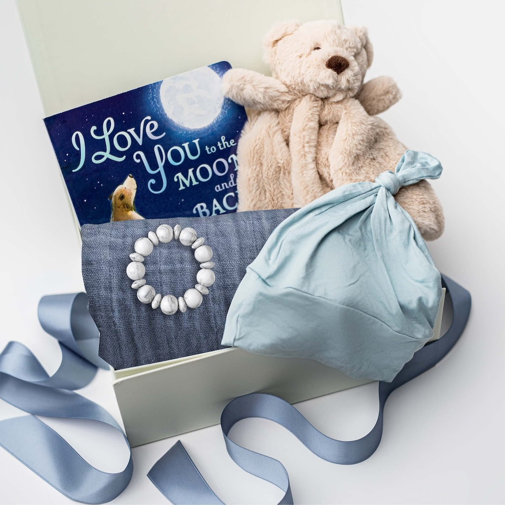 A white gift box on a white background, filled with light ivory paper shred and tied with a soft blue ribbon. Inside the box: a plush teddy bear lovey (with optional personalization), a muslin cotton swaddle blanket printed with white stars on a dusty-blue background, a soft blue stretch baby top-knot hat, a silicone and wood baby teether consisting of two wooden rings and a grey-and-white bead loop, and a board book titled “I Love You to the Moon and Back.”
