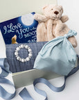 A white gift box on a white background, filled with light ivory paper shred and tied with a soft blue ribbon. Inside the box: a plush teddy bear lovey (with optional personalization), a muslin cotton swaddle blanket printed with white stars on a dusty-blue background, a soft blue stretch baby top-knot hat, a silicone and wood baby teether consisting of two wooden rings and a grey-and-white bead loop, and a board book titled “I Love You to the Moon and Back.”