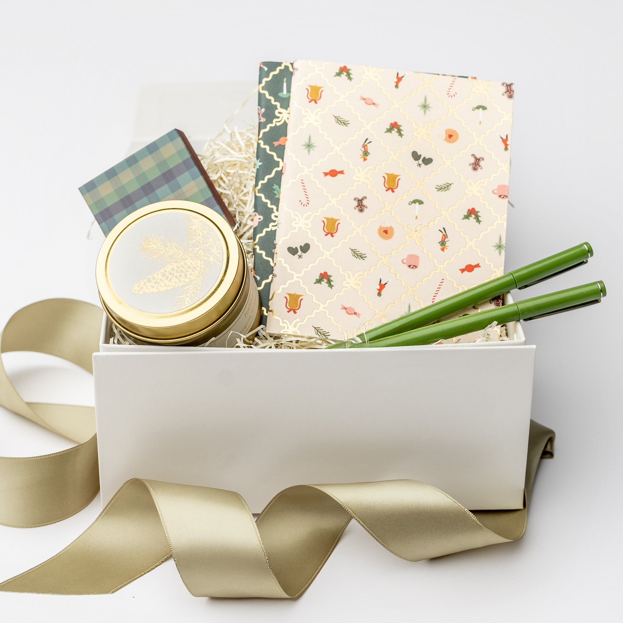 A front view of the Festive 2025 Gift Box featuring a set of two holiday argyle pocket notebooks, a set of two olive green Le Pens, a 4 oz. Spruce holiday candle, and a plaid matchbox, all arranged in a white gift box with ivory shred and tied with a green ribbon on a white background.
