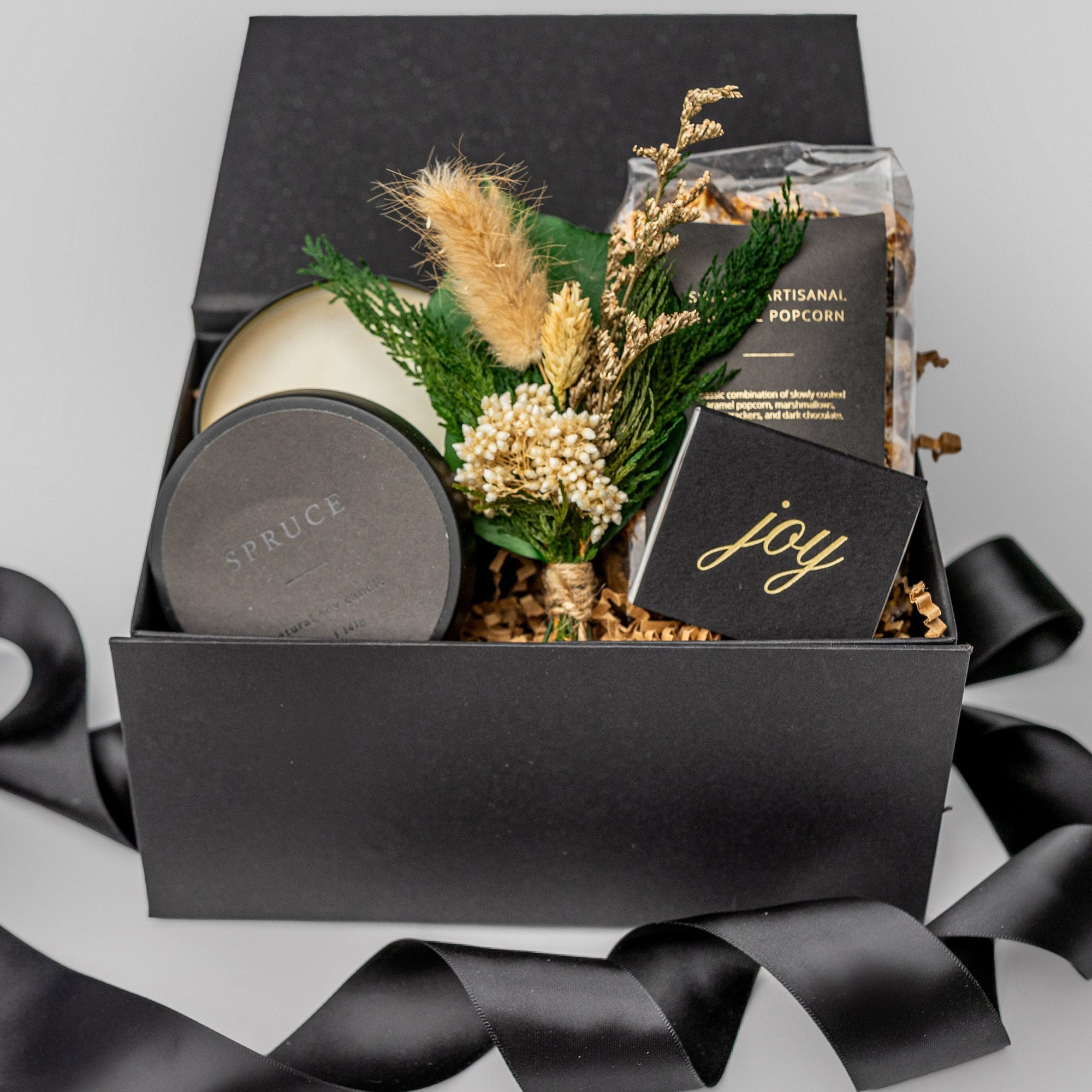 A white gift box filled with ivory paper shred on a white background. Inside the box are a bag of S’mores-flavored caramel popcorn, an 8 oz hand-poured soy candle labeled “Spruce,” a festive foil-stamped matchbox, and a handcrafted dried mini floral bouquet — a compact, holiday “Festive Favorites” gift box.