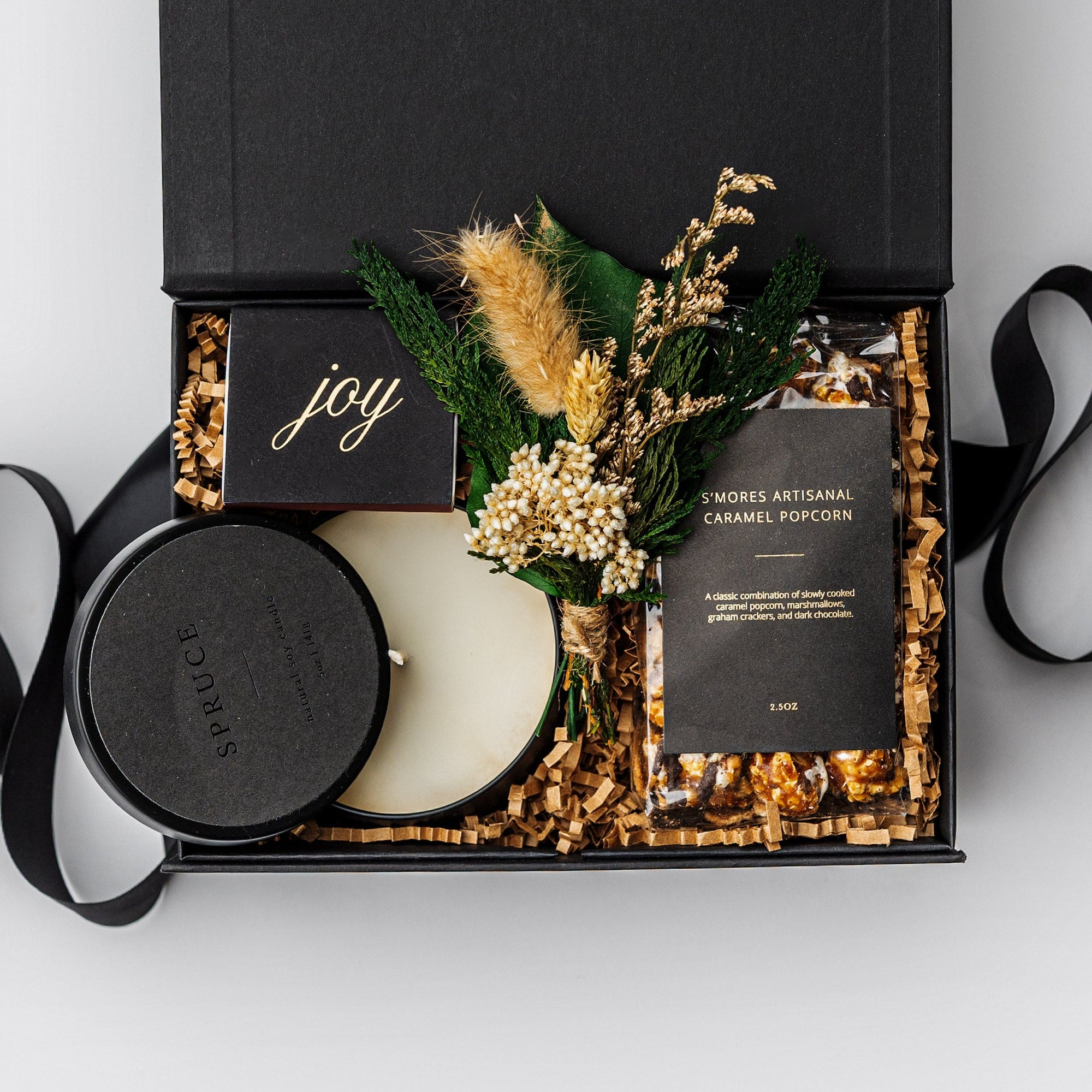 A white gift box filled with ivory paper shred on a white background. Inside the box are a bag of S’mores-flavored caramel popcorn, an 8 oz hand-poured soy candle labeled “Spruce,” a festive foil-stamped matchbox, and a handcrafted dried mini floral bouquet — a compact, holiday “Festive Favorites” gift box.
