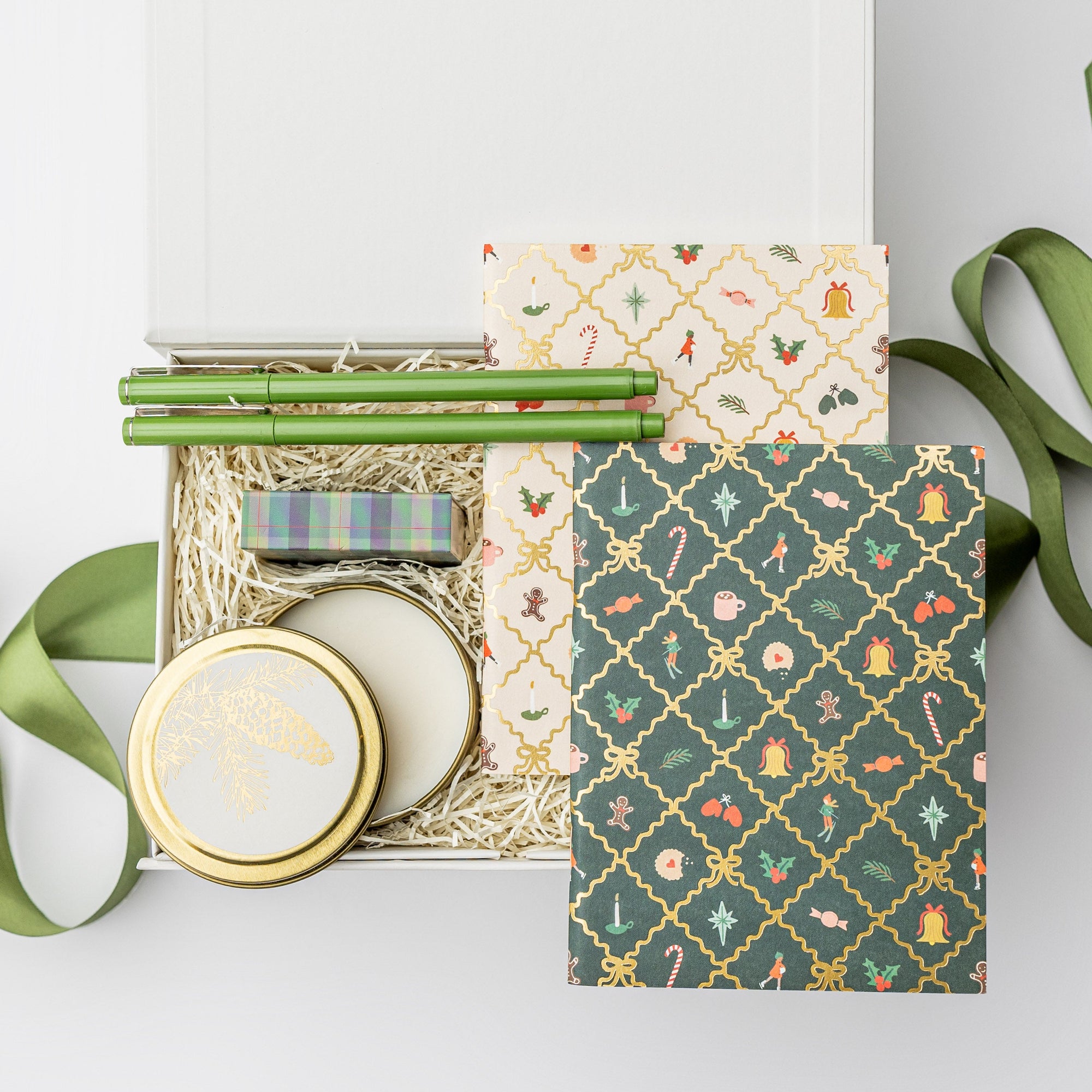 A top view of the Festive 2025 Gift Box featuring a set of two holiday argyle pocket notebooks, a set of two olive green Le Pens, a 4 oz. Spruce holiday candle, and a plaid matchbox, all arranged in a white gift box with ivory shred and tied with a green ribbon on a white background.