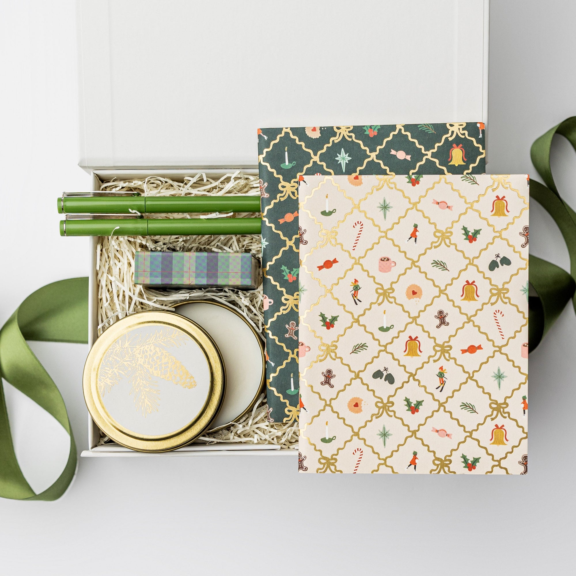 A top view of the Festive Gift Box featuring a set of two holiday argyle pocket notebooks, a set of two olive green Le Pens, a 4 oz. Spruce holiday candle, and a plaid matchbox, all arranged in a white gift box with ivory shred and tied with a green ribbon on a white background.