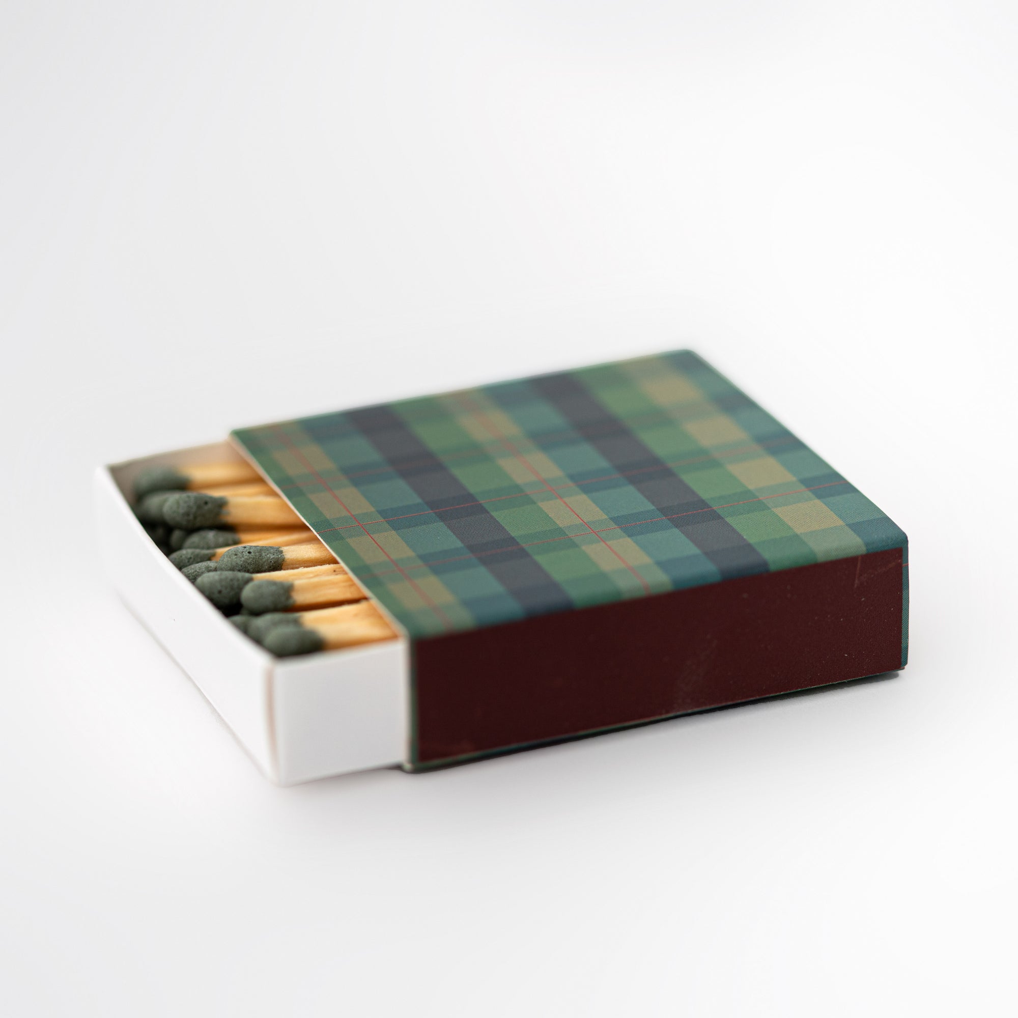 Festive plaid matchbox is featured with plaid design on the cover opened with green tip matches on a white background. 