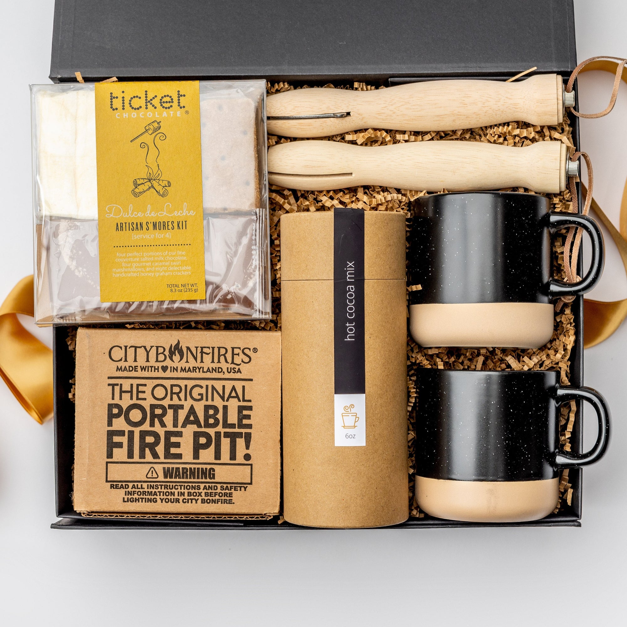A top view of the Fireside 2025 Gift Box featuring two speckled ceramic mugs (black & tan bottom), a jar of hot cocoa mix, one dulce de leche s'mores kit, a portable fire pit, and a pair of retractable skewers, all arranged in a white gift box with brown shred and tied with an orange ribbon on a white background.