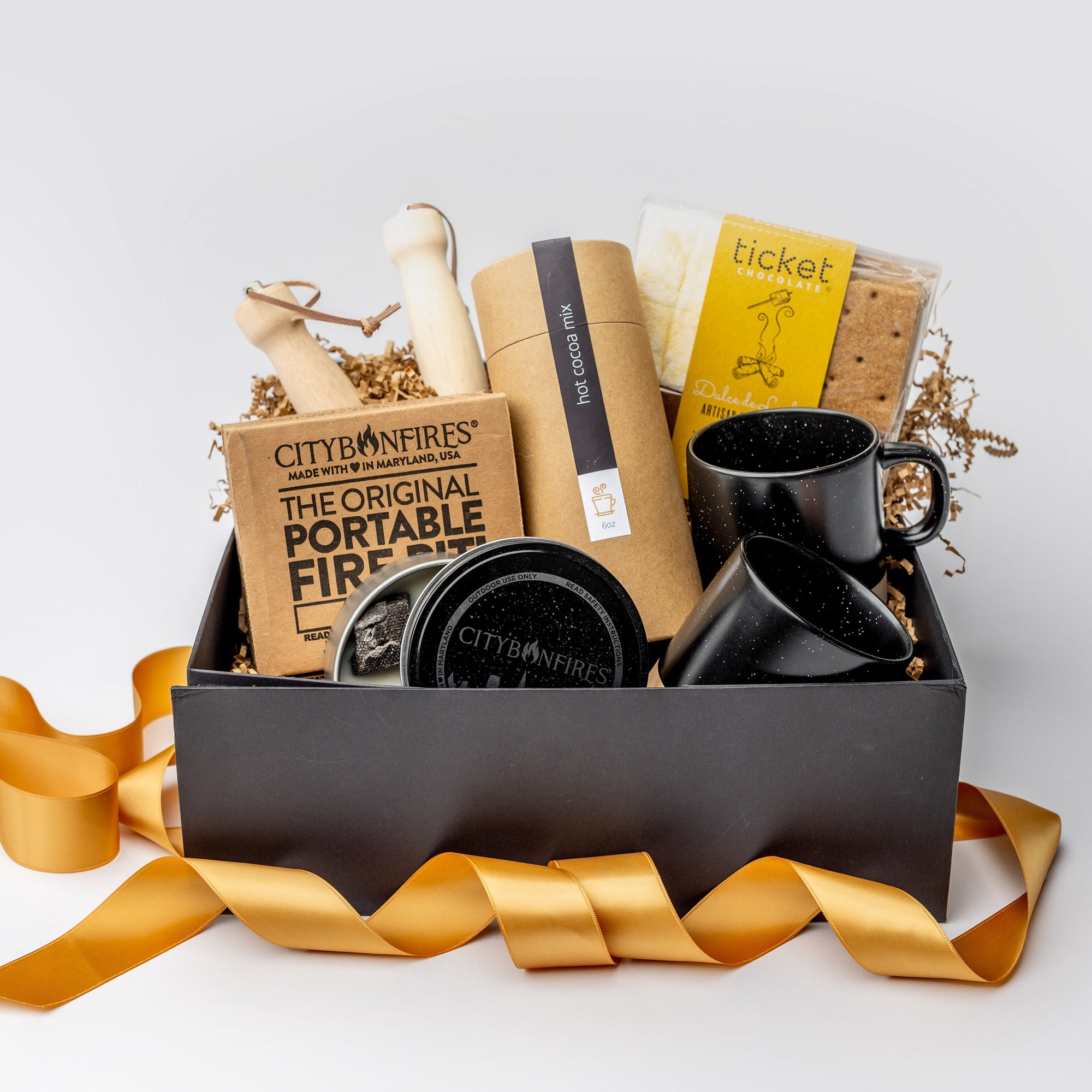 A front view of the Fireside 2025 Gift Box featuring two speckled ceramic mugs (black & tan bottom), a jar of hot cocoa mix, one dulce de leche s'mores kit, a portable fire pit, and a pair of retractable skewers, all arranged in a white gift box with brown shred and tied with an orange ribbon on a white background.