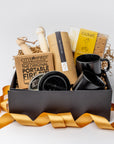 A front view of the Fireside 2025 Gift Box featuring two speckled ceramic mugs (black & tan bottom), a jar of hot cocoa mix, one dulce de leche s'mores kit, a portable fire pit, and a pair of retractable skewers, all arranged in a white gift box with brown shred and tied with an orange ribbon on a white background.