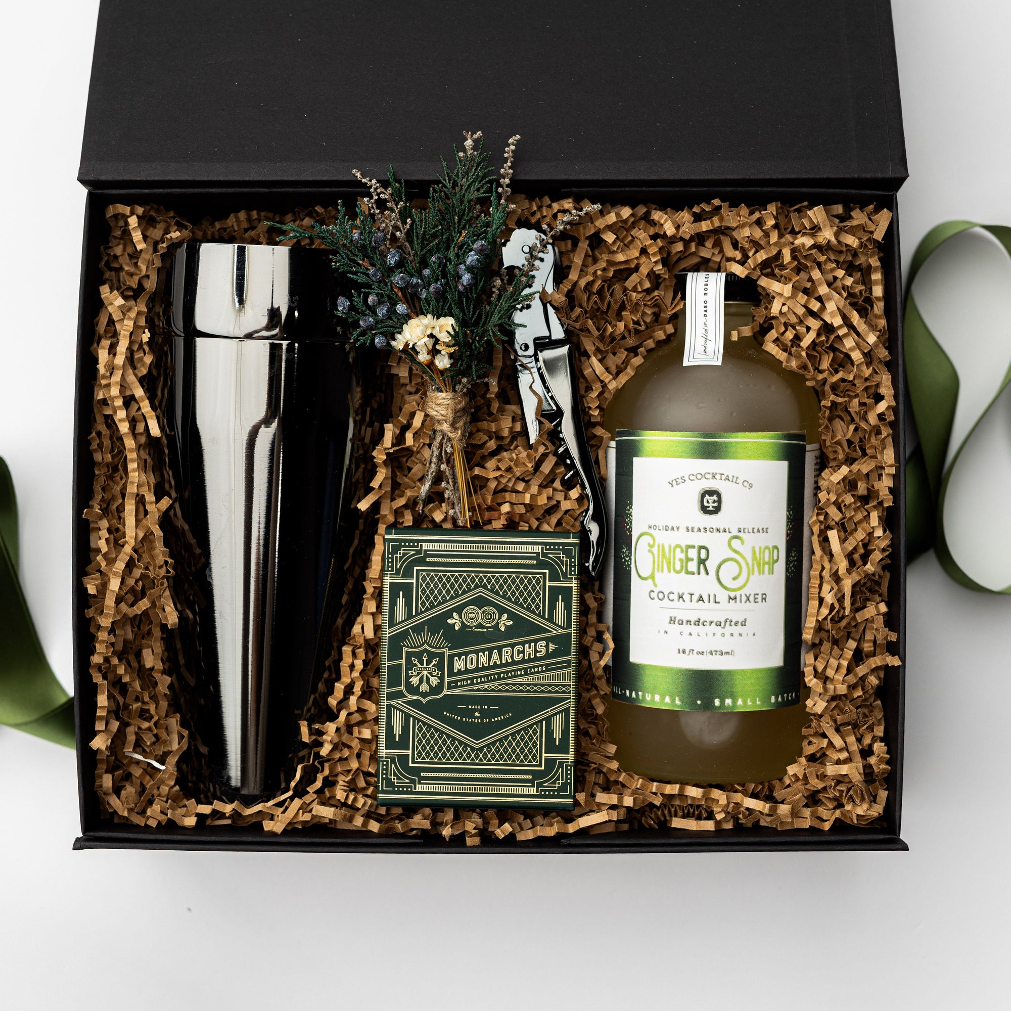 Fireside Spirits Gift Box is featured with a black box and a green ribbon underneath. Inside the box, on the left, there is a gunmetal-plated cocktail shaker. In the middle, a floral bundle, a gunmetal-plated corkscrew, and Monarch luxury playing cards with green and gold details. On the right, a Ginger Snap cocktail mixer with a white label and green details. All items are arranged in front of brown shred in the back, displayed on a white background.