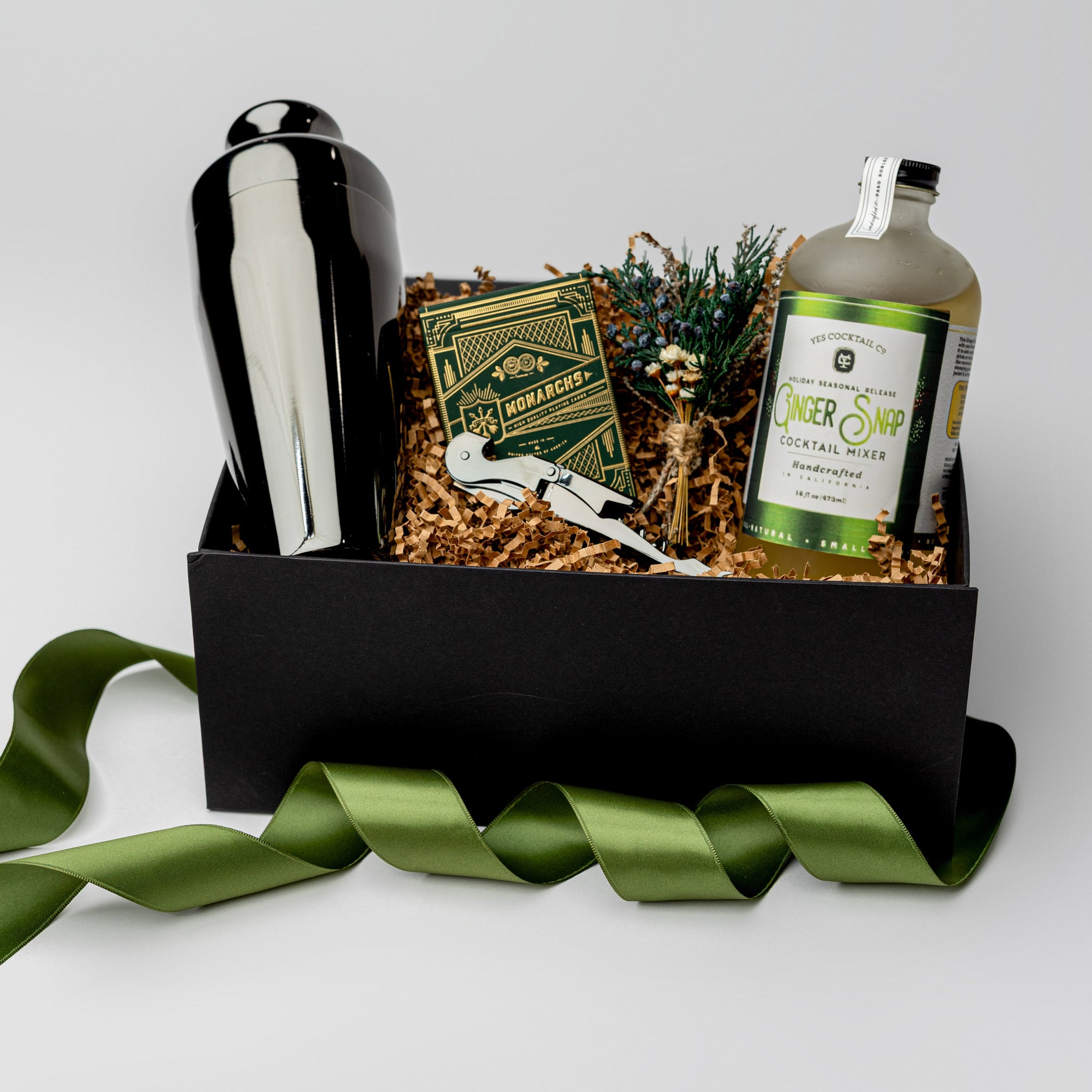 Fireside Spirits Gift Box is featured with a black box and a green ribbon around it. Inside the box, on the left, there is a gunmetal-plated cocktail shaker. In the middle, a floral bundle, a gunmetal-plated corkscrew, and Monarch luxury playing cards with green and gold details. On the right, a Ginger Snap cocktail mixer with a white label and green details. All items are arranged in front of brown shred in the back, displayed on a white background.