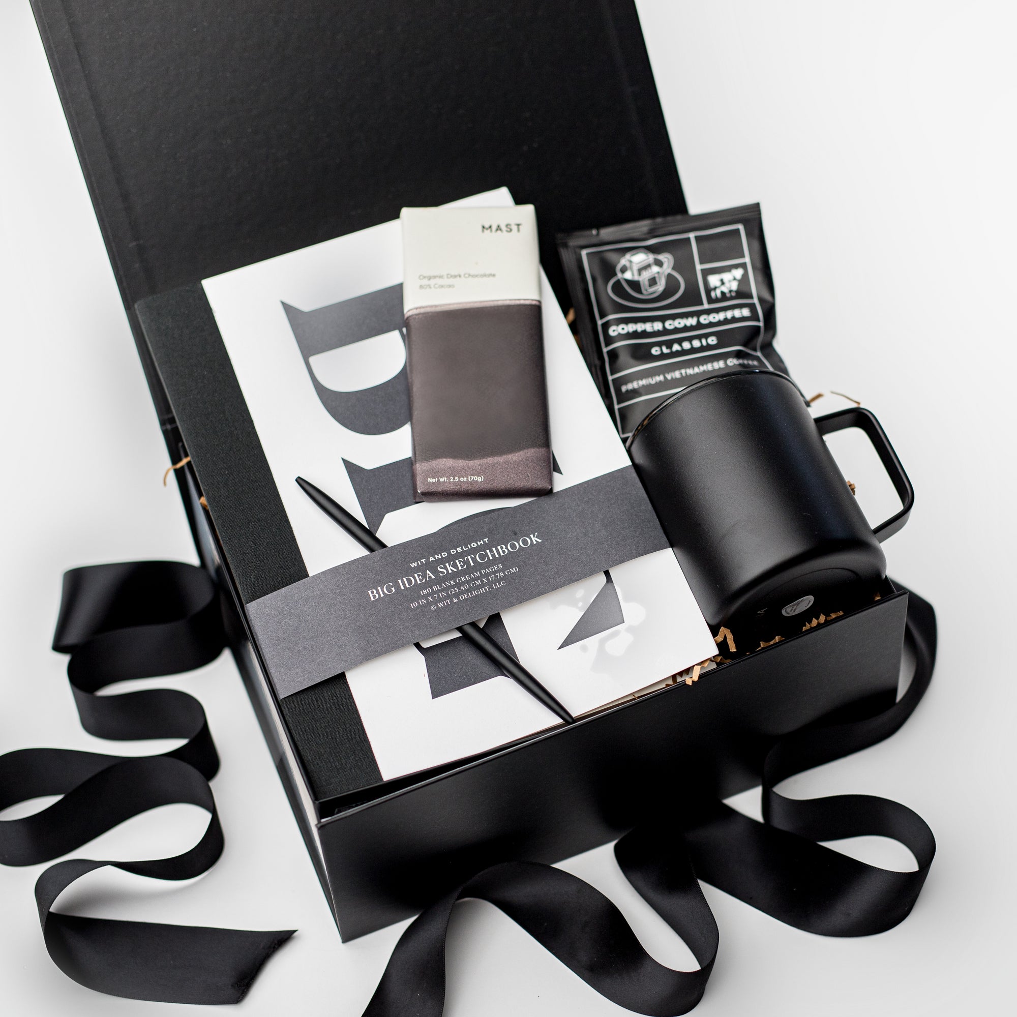 A top view of the Seize the Day Gift Box presented in a black gift box filled with brown shred and tied with a black ribbon, featuring a Big Idea sketchbook, a minimalist black bullet pen, a 12 oz. black vacuum insulated camp mug, a Copper Cow single-serve pour-over coffee, and a MAST organic dark chocolate bar (80% cacao), all arranged on a white background.