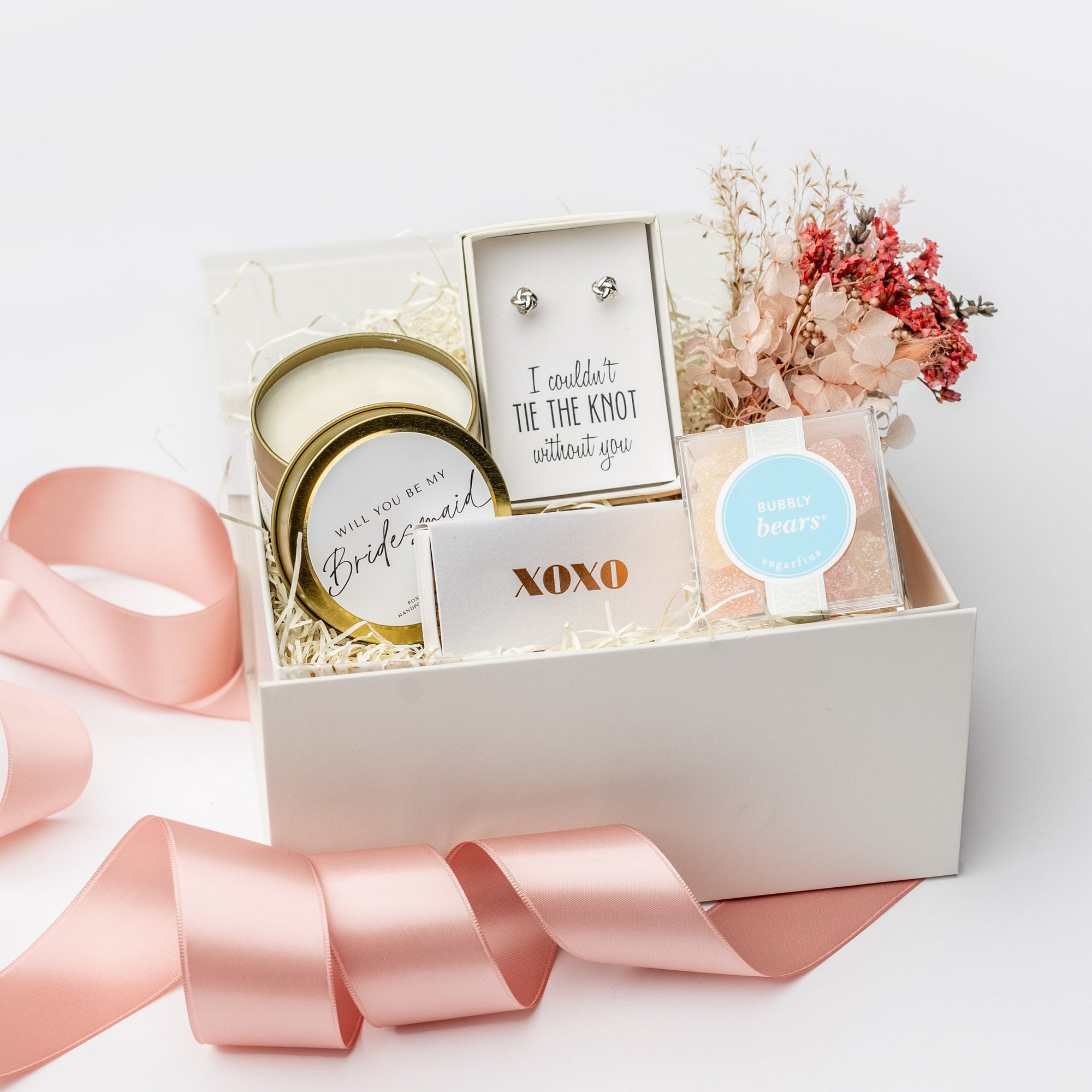 The Be My Bridesmaid Gift Box is featured in a top-side view of our signature white gift box filled with ivory shred. On the left is a “Will You Be My Bridesmaid?” candle in a gold tin, in the center a metallic rose gold “XOXO” matchbox and “I Couldn’t Tie the Knot Without You” earrings, and on the right a dried floral bundle and champagne bubbly gummy bears. Displayed on a white background with a light pink ribbon surrounding it.