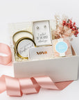 The Be My Bridesmaid Gift Box is featured in a top-side view of our signature white gift box filled with ivory shred. On the left is a “Will You Be My Bridesmaid?” candle in a gold tin, in the center a metallic rose gold “XOXO” matchbox and “I Couldn’t Tie the Knot Without You” earrings, and on the right a dried floral bundle and champagne bubbly gummy bears. Displayed on a white background with a light pink ribbon surrounding it.