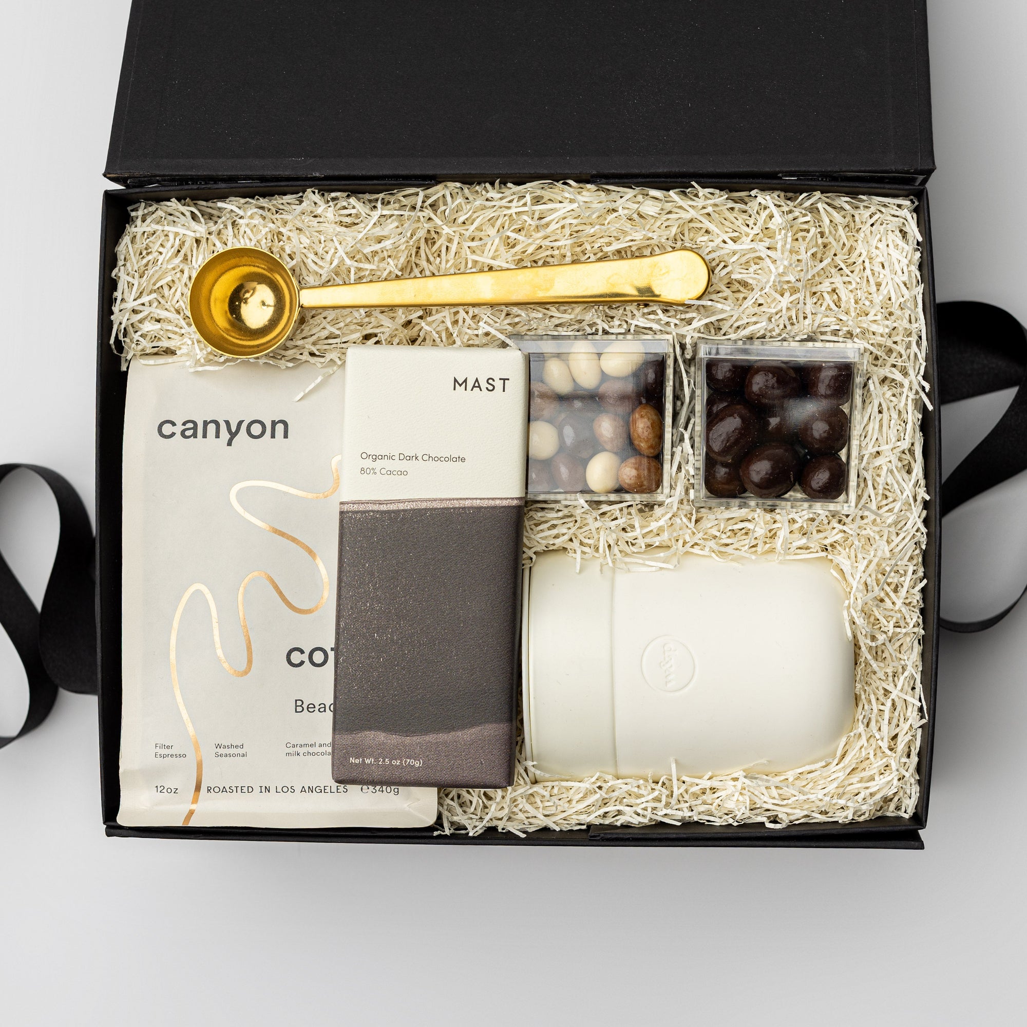 A top view of our Brew Gift Box is featured in a black gift box filled with ivory shred. On top is a gold coffee scoop, and underneath, from left to right, Canyon Beachwood Coffee, classic dark chocolate cold brew cordials, chocolate-covered espresso beans, and a cream ceramic travel mug with a silicone sleeve. Displayed on a white background with a black ribbon crossing underneath the box.
