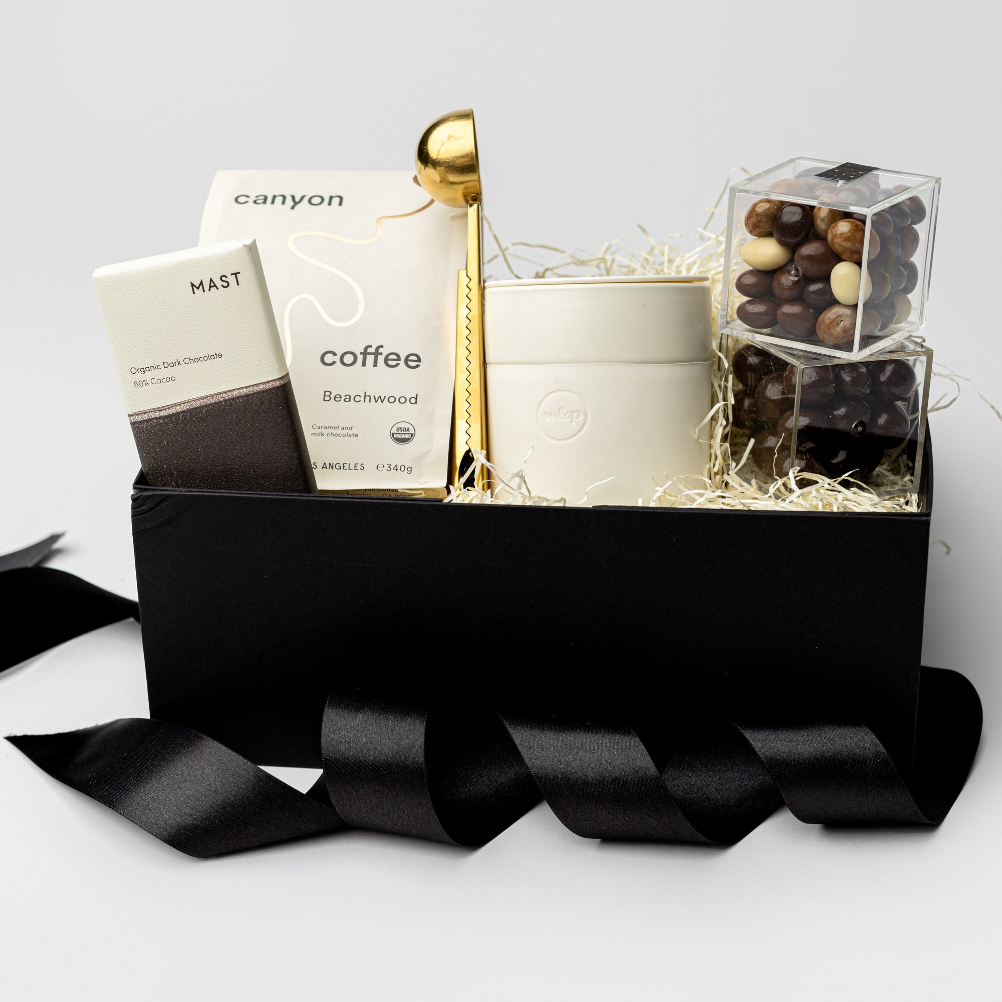A front view of our Brew Gift Box is featured in a black gift box filled with ivory shred. From left to right, it includes chocolate coffee, a gold coffee scoop and cream ceramic tumbler with a silicone sleeve in the center, and on the right, classic dark chocolate cold brew cordials and chocolate-covered espresso beans. Displayed on a white background with a black ribbon crossing underneath the box.